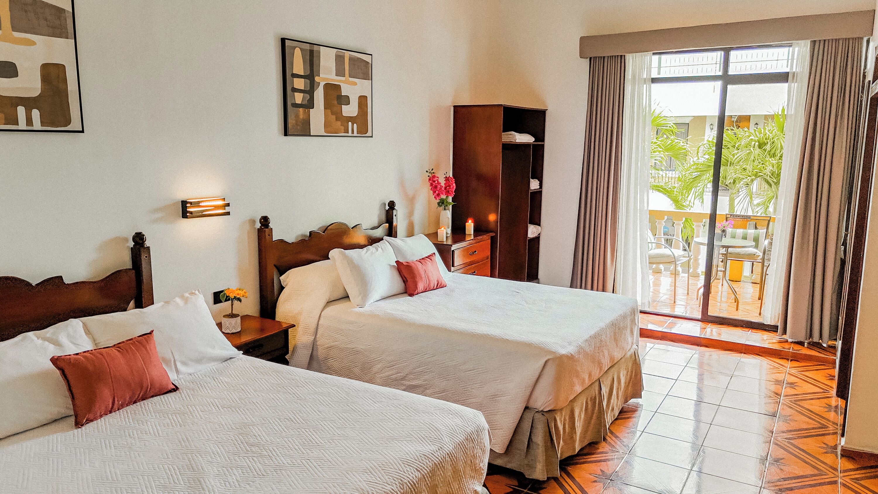 Habitación de hotel con dos camas, una mesita de noche, una lámpara de pared, una ventana con cortinas y vistas al exterior.