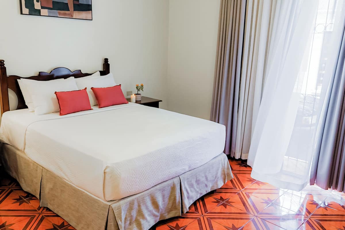 A hotel room with a bed, a nightstand, a vase with flowers, and a window with curtains.