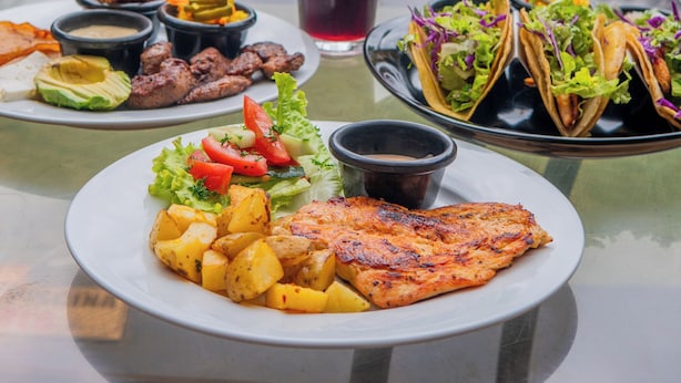 Une table à manger dressée avec une variété de plats, notamment du poisson grillé, des tacos et une salade, le tout disposé devant une vue sur la piscine.