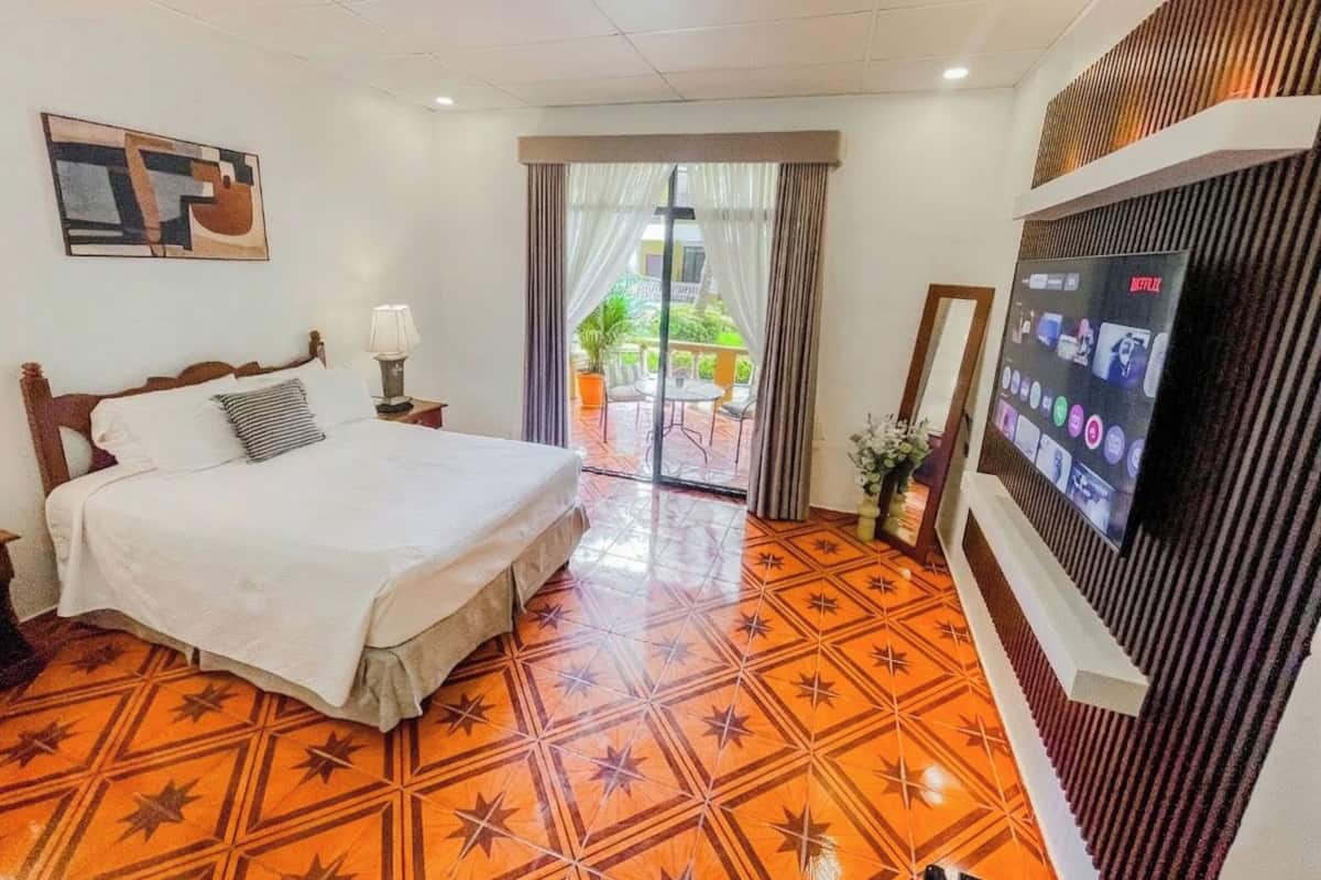 A hotel room with a bed, a television, a patterned floor, and a view of the outdoors.