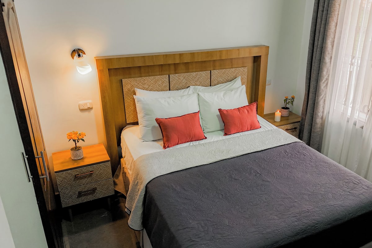 A neatly made bed with a wooden headboard, a bedside table with a lamp, and a nightstand with a flower vase.