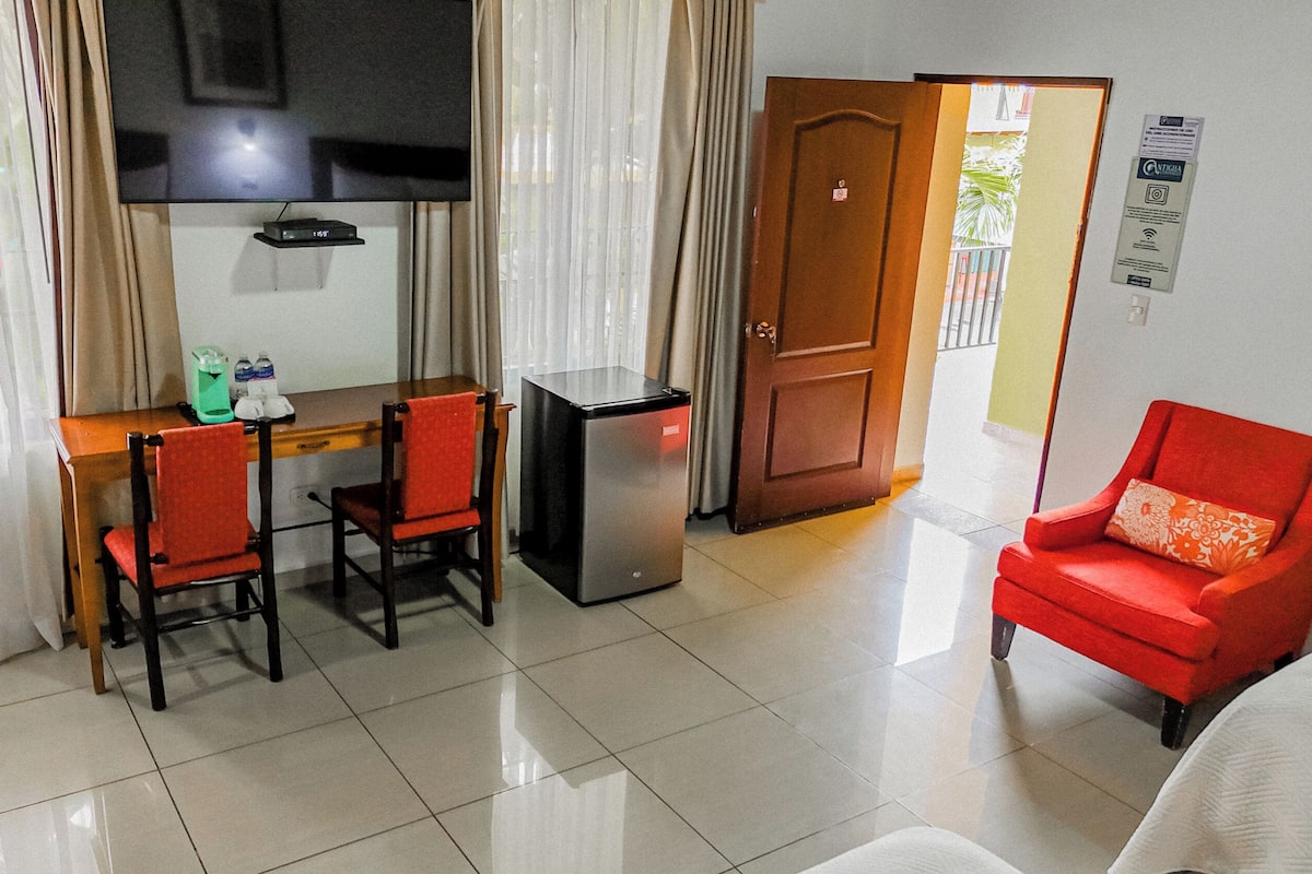 A compact hotel room with a dining area, a compact refrigerator, a red armchair, and a door leading to an outdoor area.