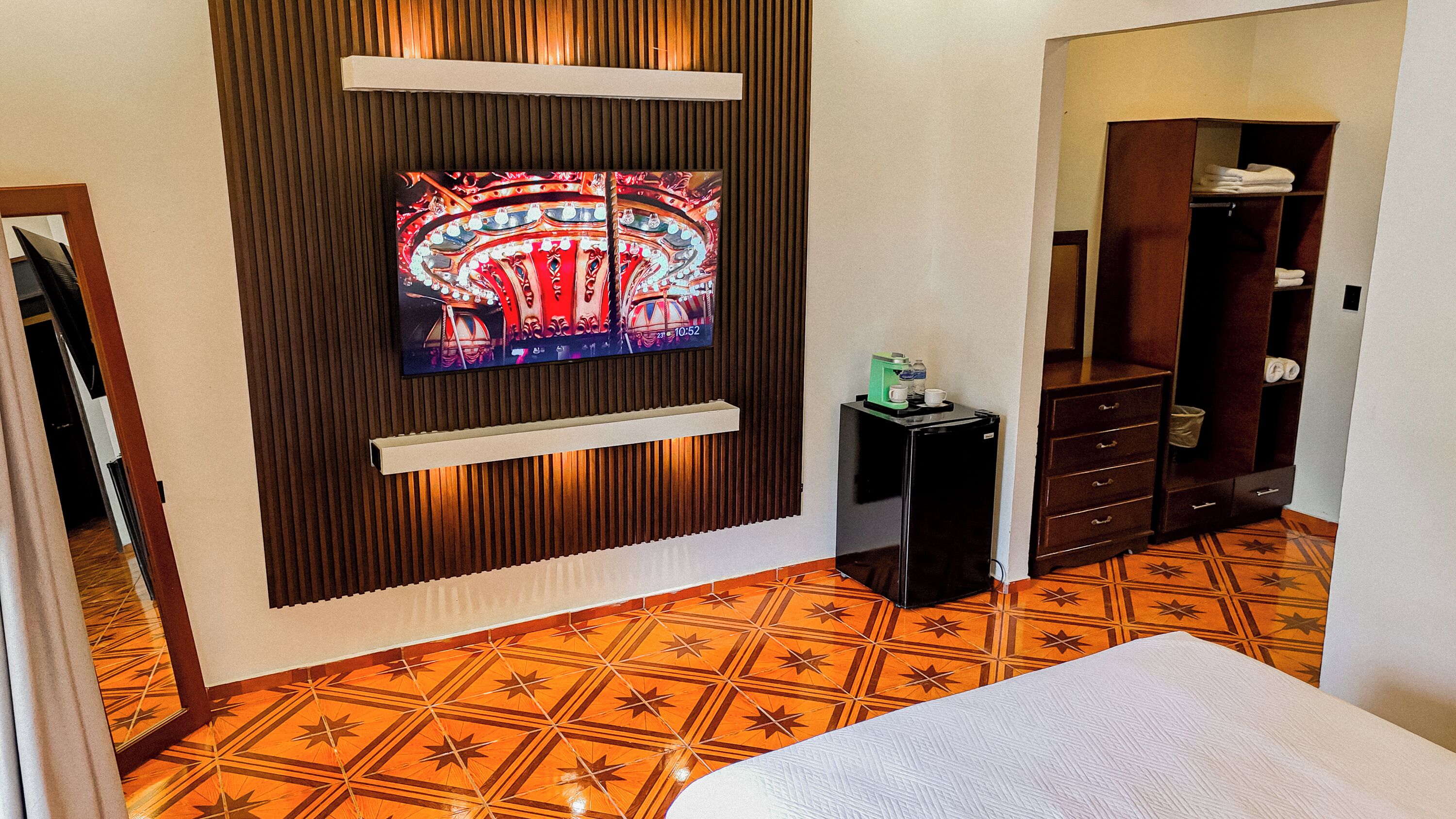 A hotel room with a television mounted on the wall, a minibar, a wardrobe, and a patterned floor.
