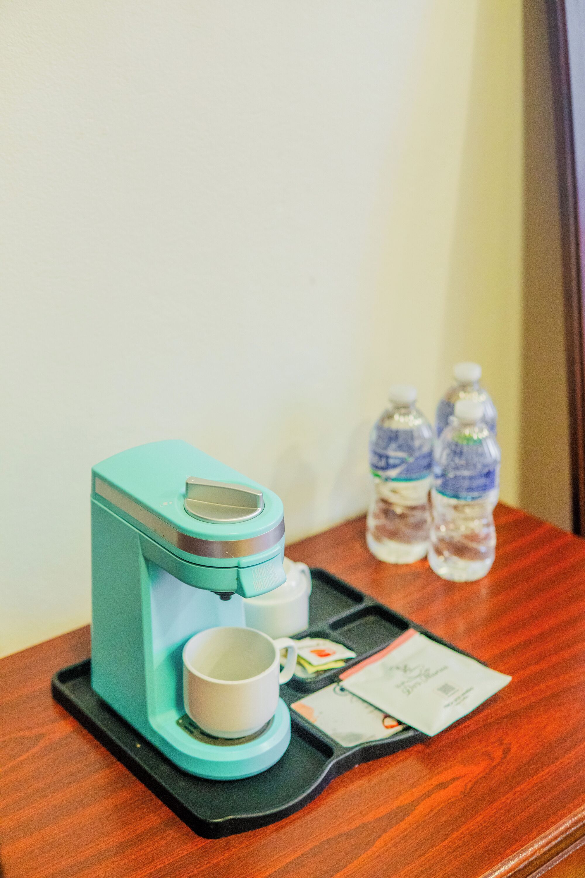 Una cafetera color turquesa sobre una bandeja con una taza blanca y un sobre pequeño, dos botellas de agua y un recibo sobre una mesa de madera.