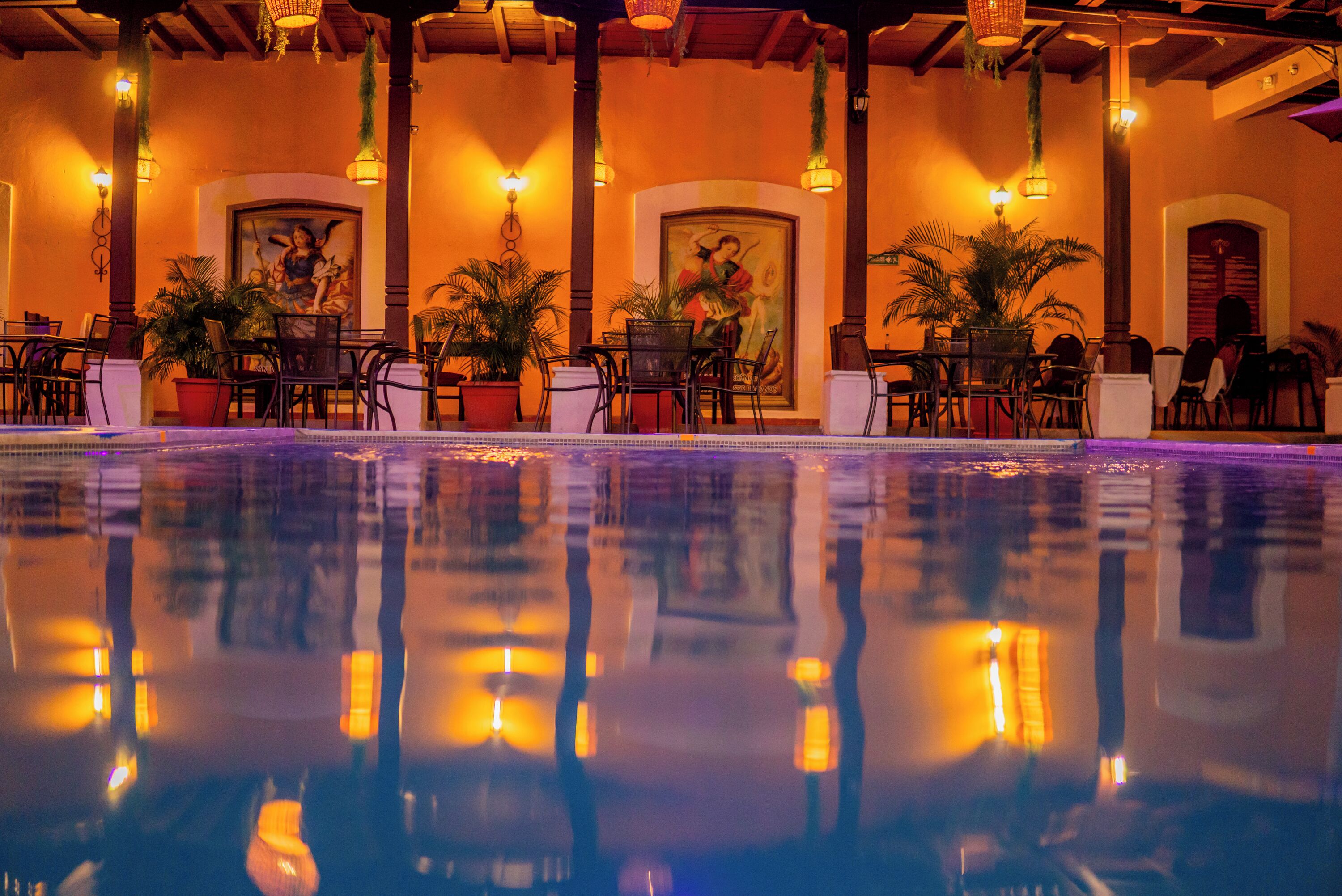 A poolside area with tables and chairs, potted plants, and wall-mounted lights.