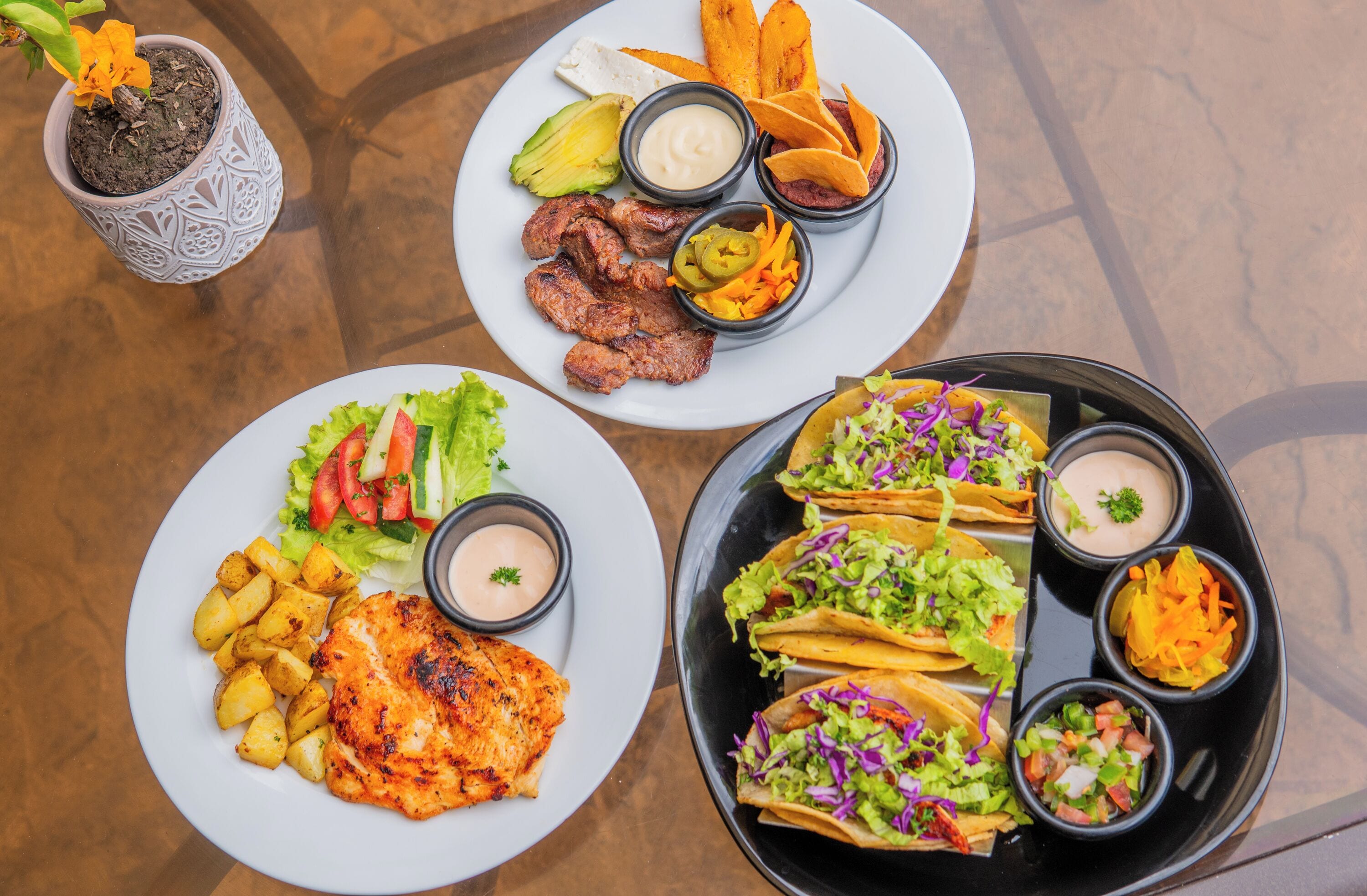 A table with three plates of food, including tacos, grilled meat, and potatoes.