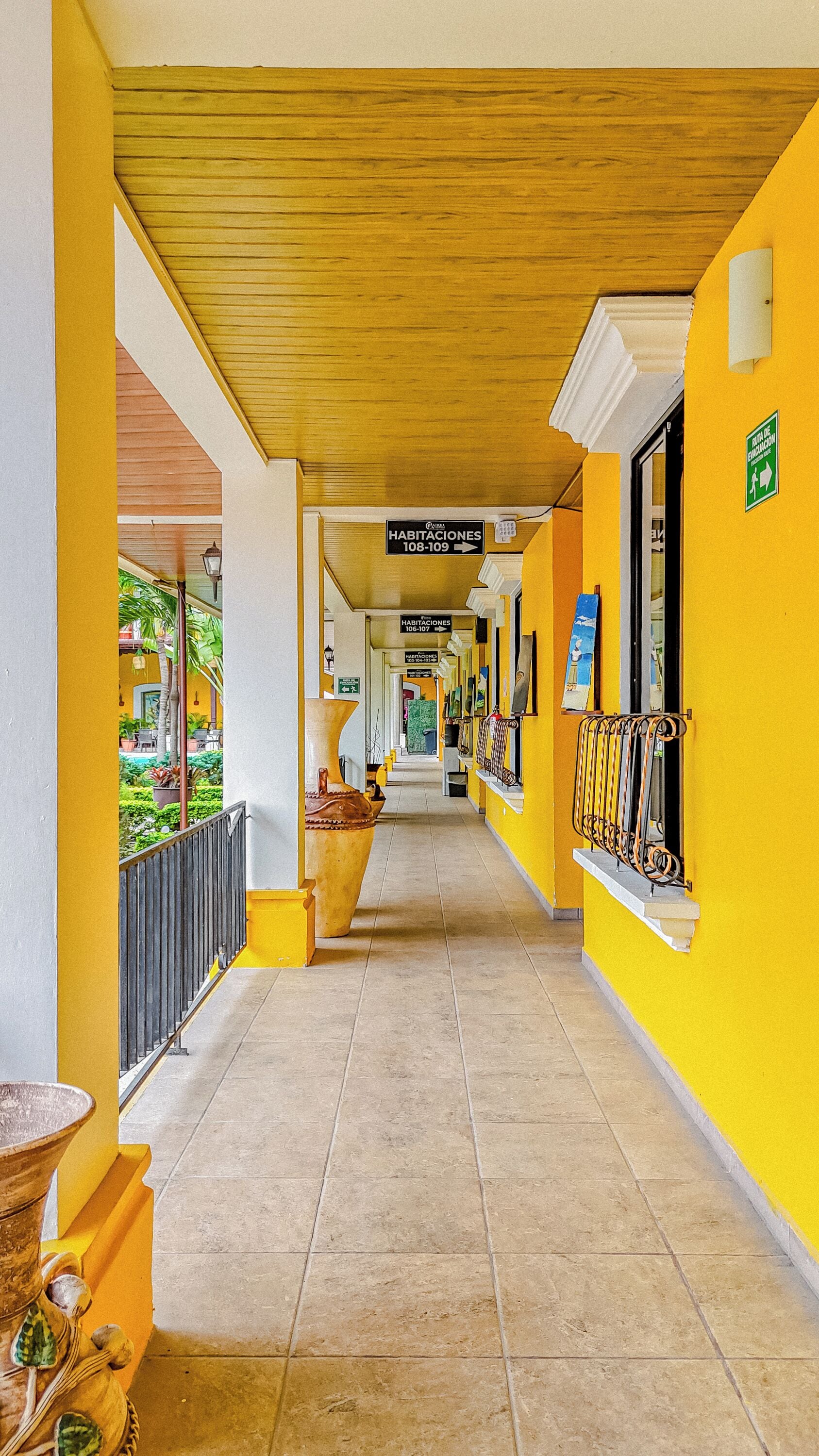 A corridor with yellow walls, a tiled floor, and signs indicating room numbers.