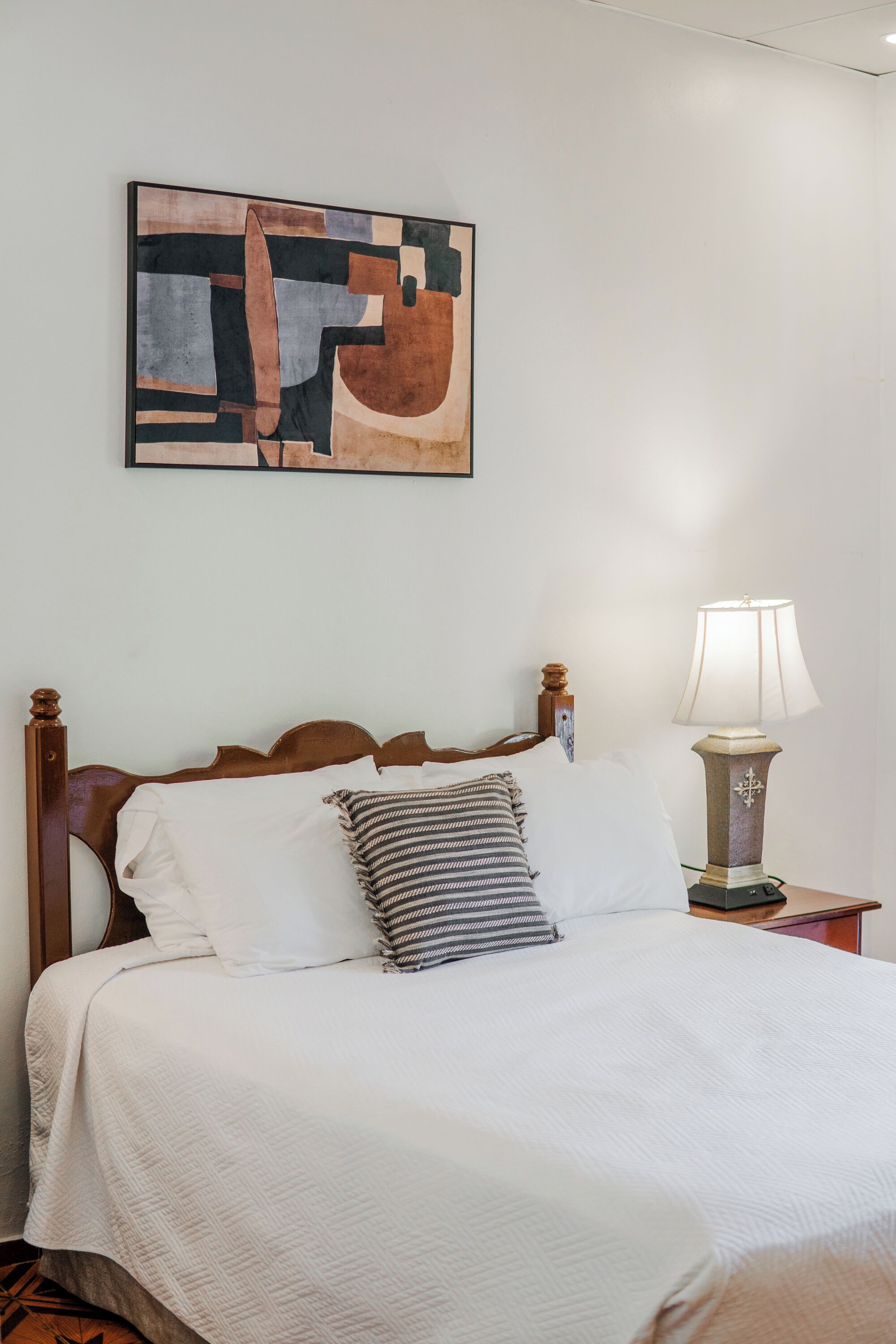 A neatly made bed with a white comforter and striped pillow, a wooden headboard, a bedside lamp, and a wall-mounted abstract painting.
