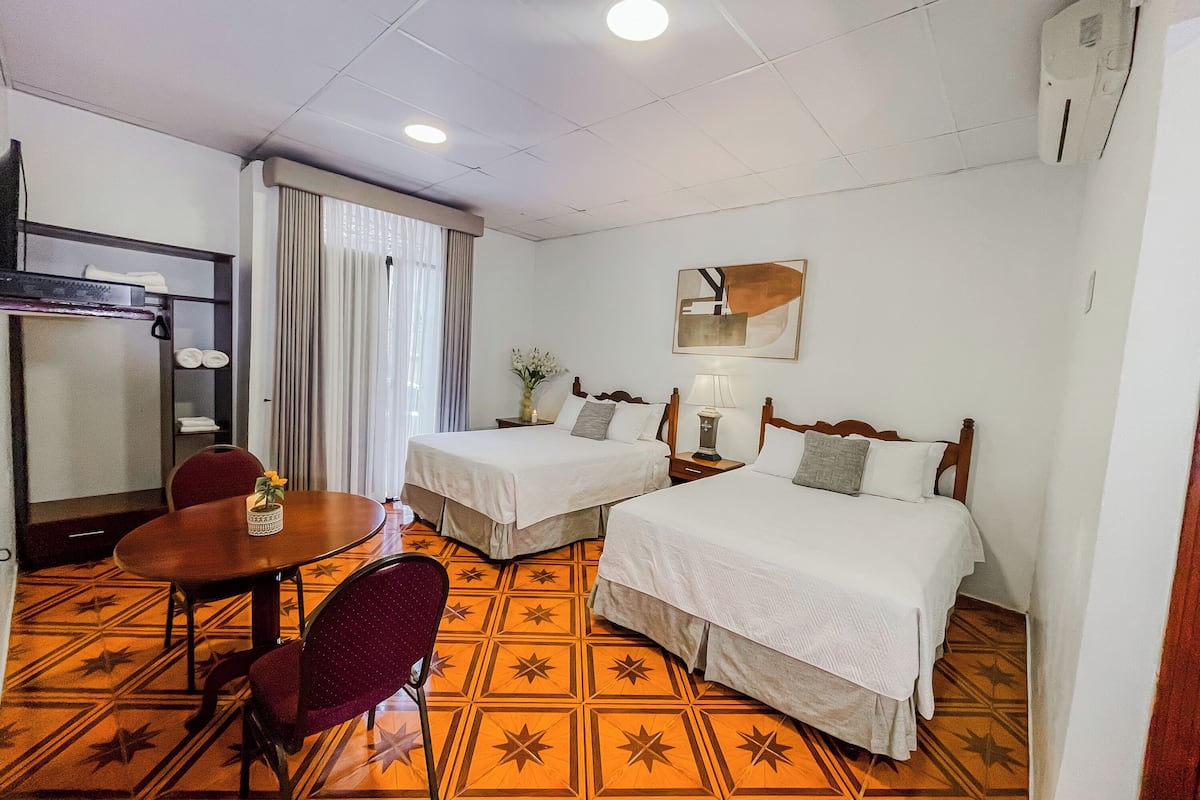 A hotel room with two beds, a dining table, a chair, a lamp, and a painting on the wall.