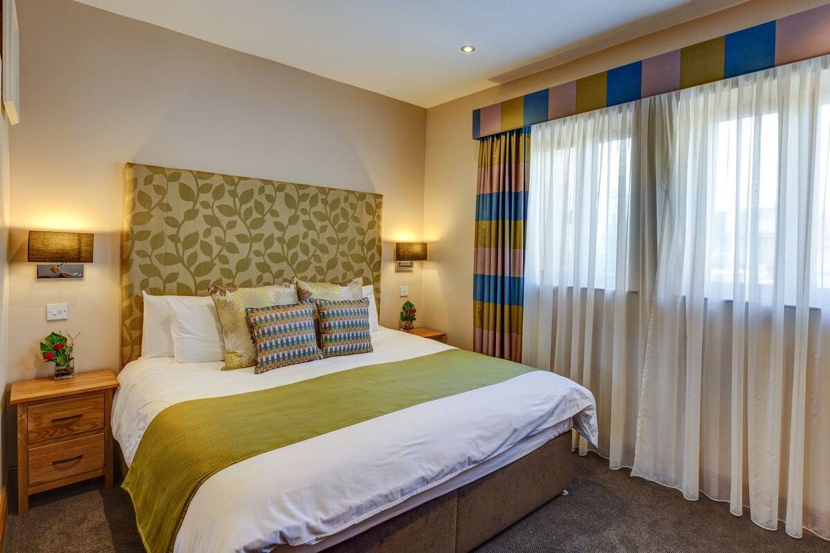 Un dormitorio ordenado con una cama grande, dos mesitas de noche y una ventana con cortinas.