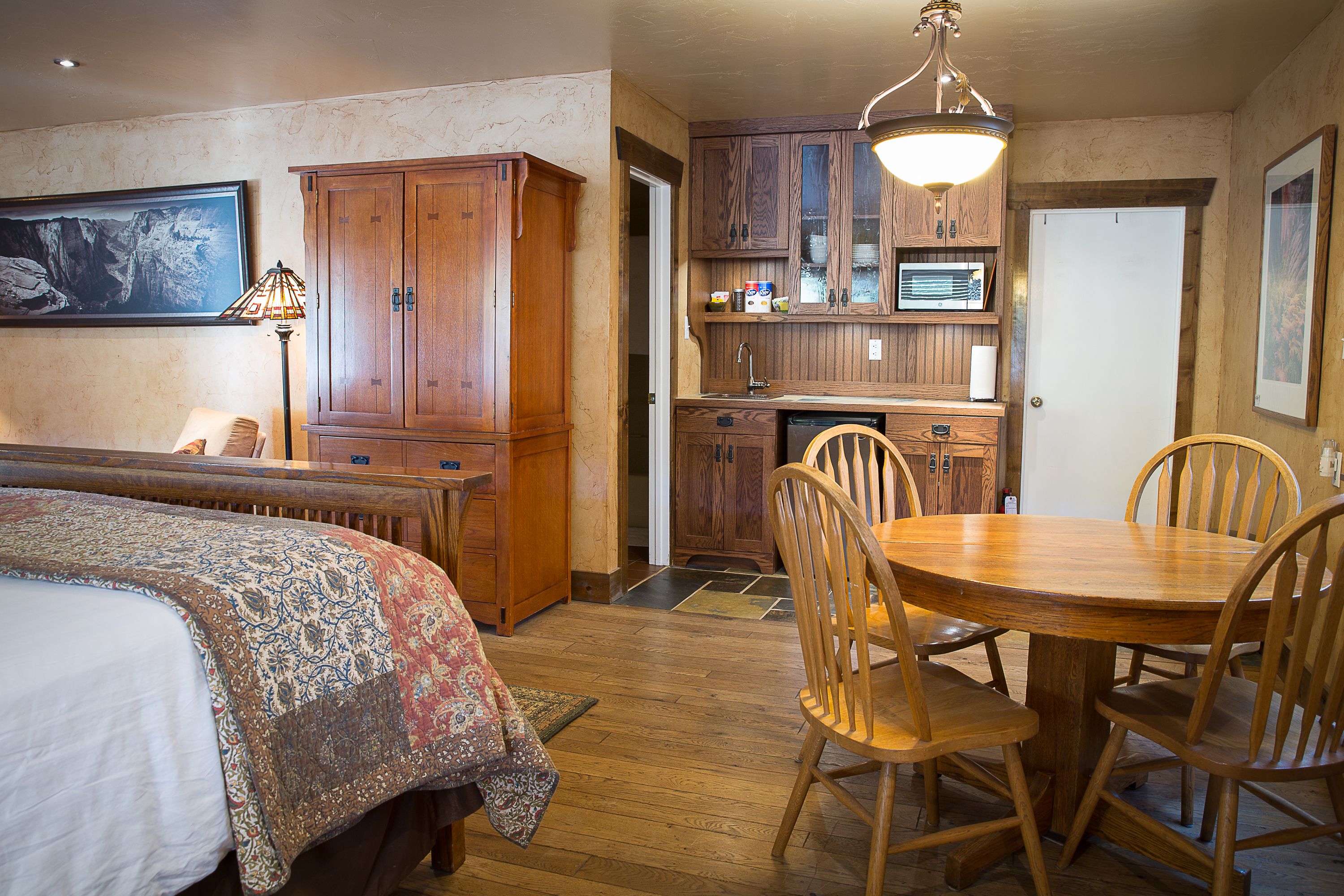 Una habitación acogedora con una cama, una mesa de comedor con sillas, una zona de cocina con armarios de madera y un cuadro grande en la pared.