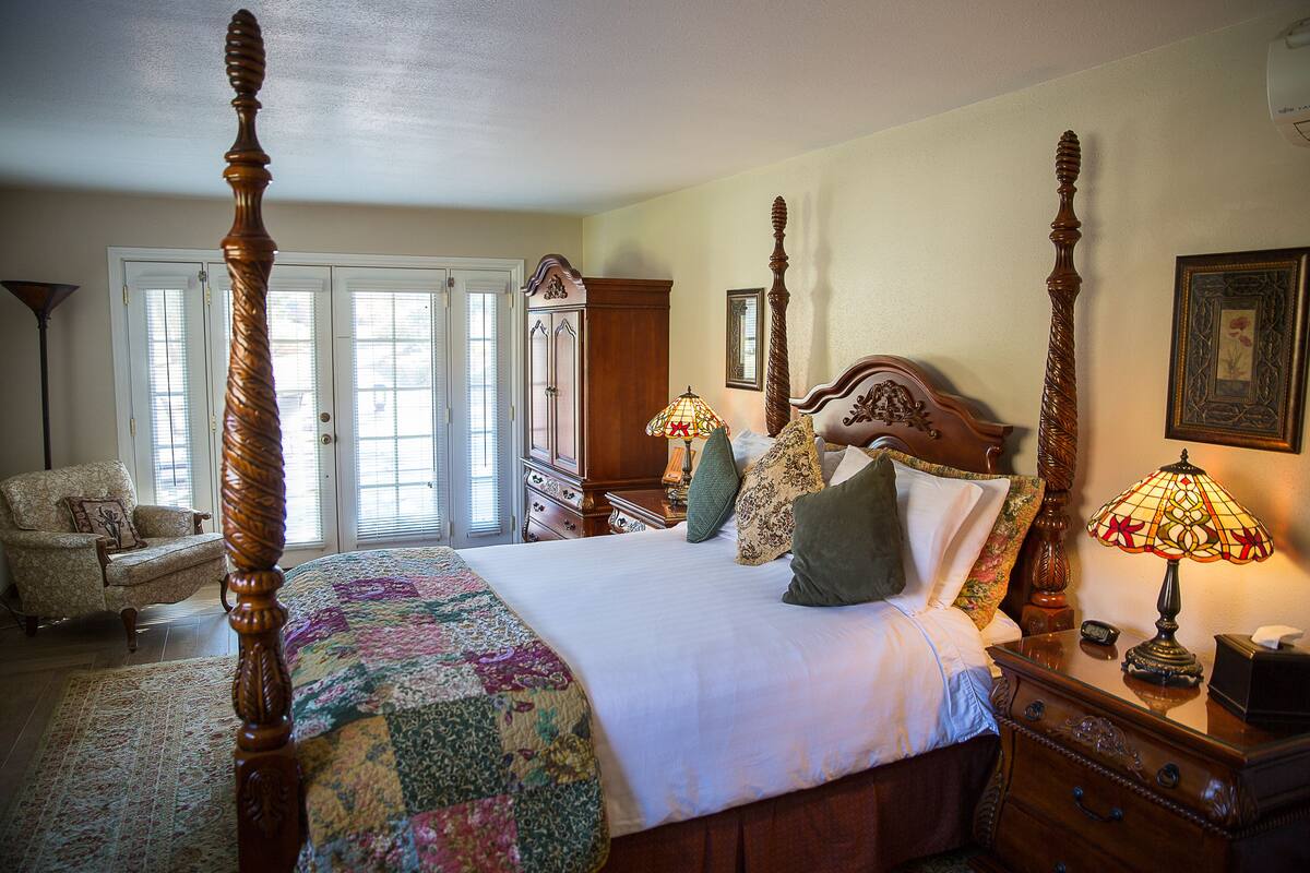 A four-poster bed with a quilt, a wooden nightstand with a lamp, a chair, and a dresser in a well-lit room with large windows.