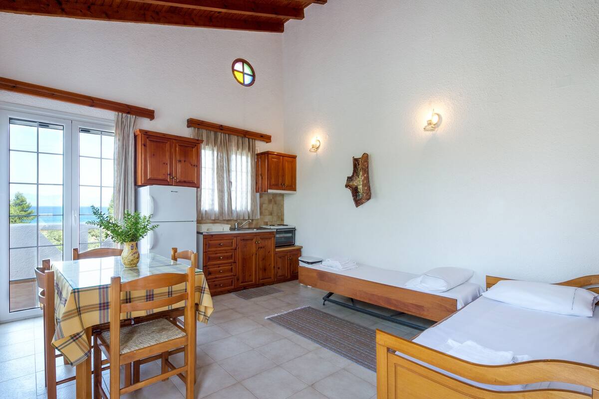 A compact kitchen with wooden cabinets, a dining table with chairs, a small refrigerator, and a bench with pillows.