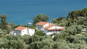 A coastal property with two white buildings, red-tiled roofs, surrounded by dense greenery and a view of the sea.