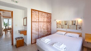 A hotel room with a bed, a wooden cabinet, a mirror, a wall-mounted light, and a door with a green exit sign.
