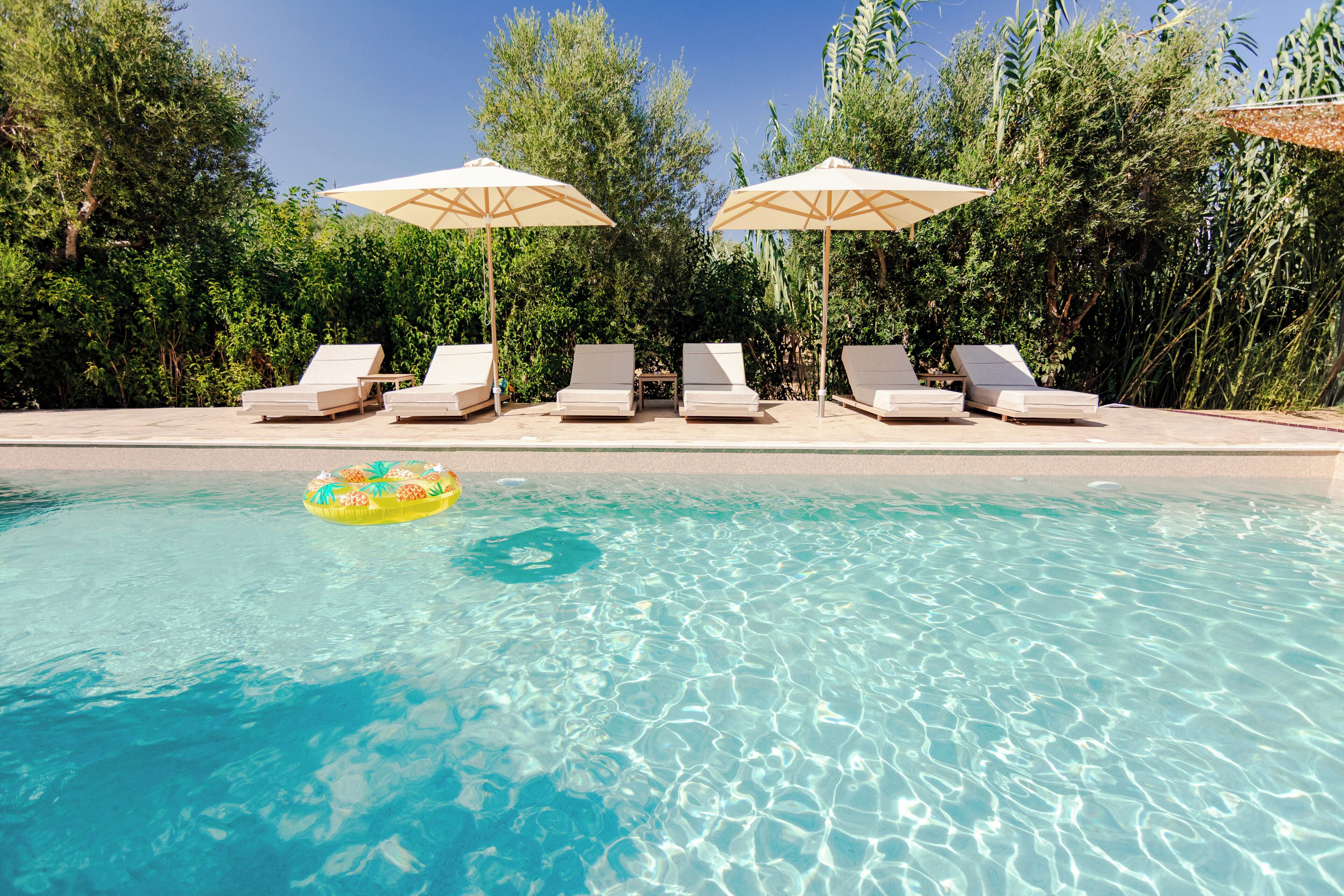 A clear pool with six lounge chairs and two umbrellas, surrounded by dense greenery.