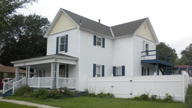A two-story house with a front porch, a staircase leading to a second-floor balcony, and a fenced yard. - Hillside Cottages (Missouri Valley)