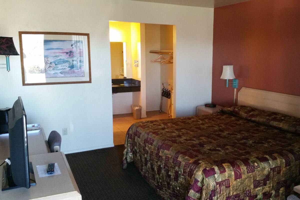 Habitación de hotel con cama, escritorio, silla, televisor, un cuadro, baño y lámpara.