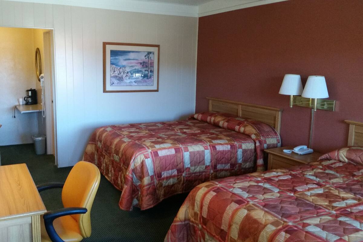Habitación de hotel con dos camas, un escritorio con televisor, una silla, un cuadro en la pared y una lámpara.