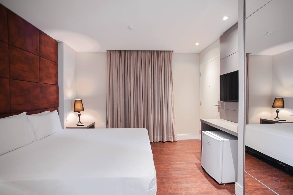 A hotel room with a large bed, a desk with a TV, a mini-fridge, and a wardrobe.