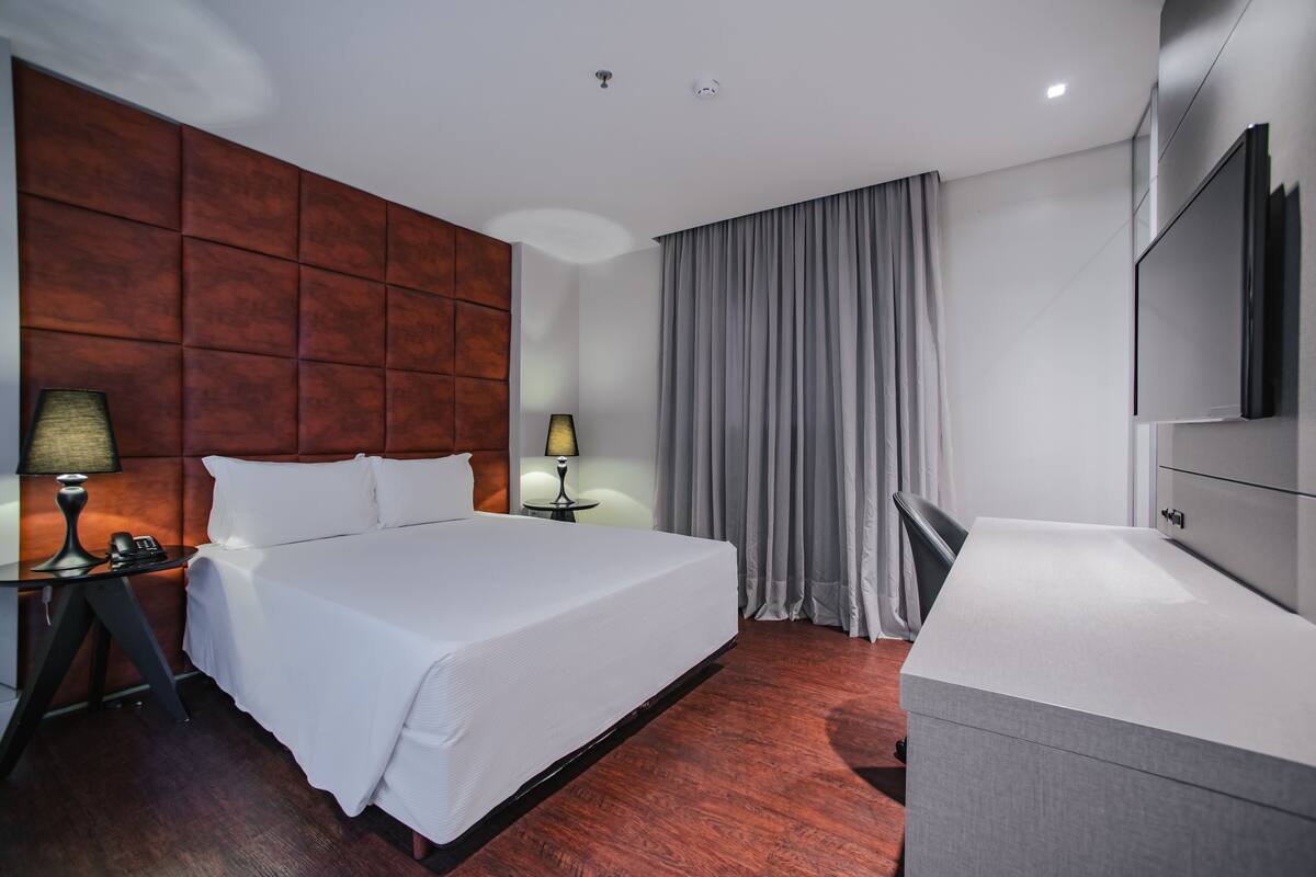 A hotel room with a large bed, a desk with a lamp, a TV, and a grey curtain.