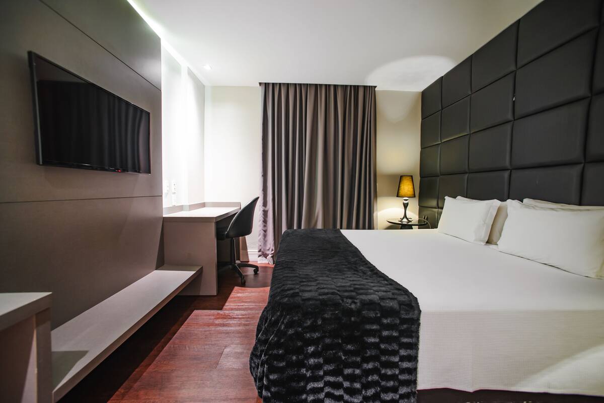 A hotel room with a bed, a desk, a chair, a TV mounted on the wall, and a window with curtains.