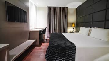 A hotel room with a bed, a desk, a chair, a TV mounted on the wall, and a window with curtains.