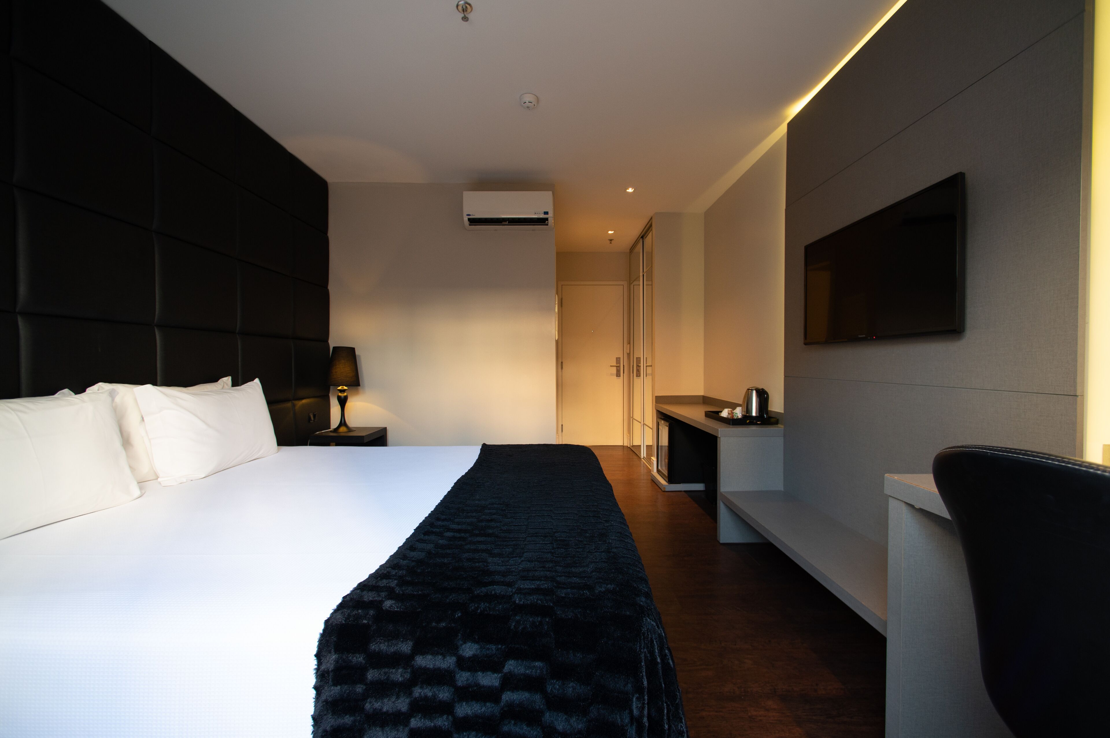 A modern hotel room with a large bed, a flat-screen TV mounted on the wall, a bedside table with a lamp, and a small kitchenette area.