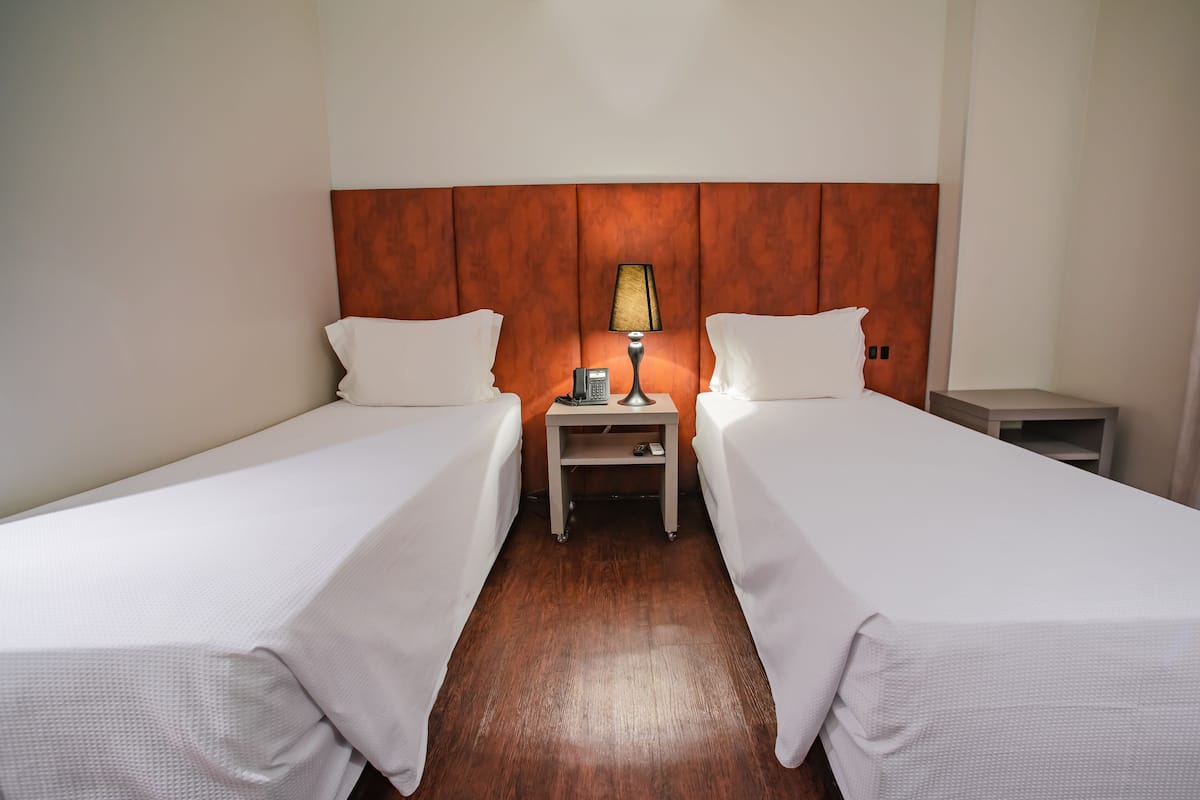 A hotel room with two single beds, a wooden headboard, a nightstand with a lamp, and a small table.