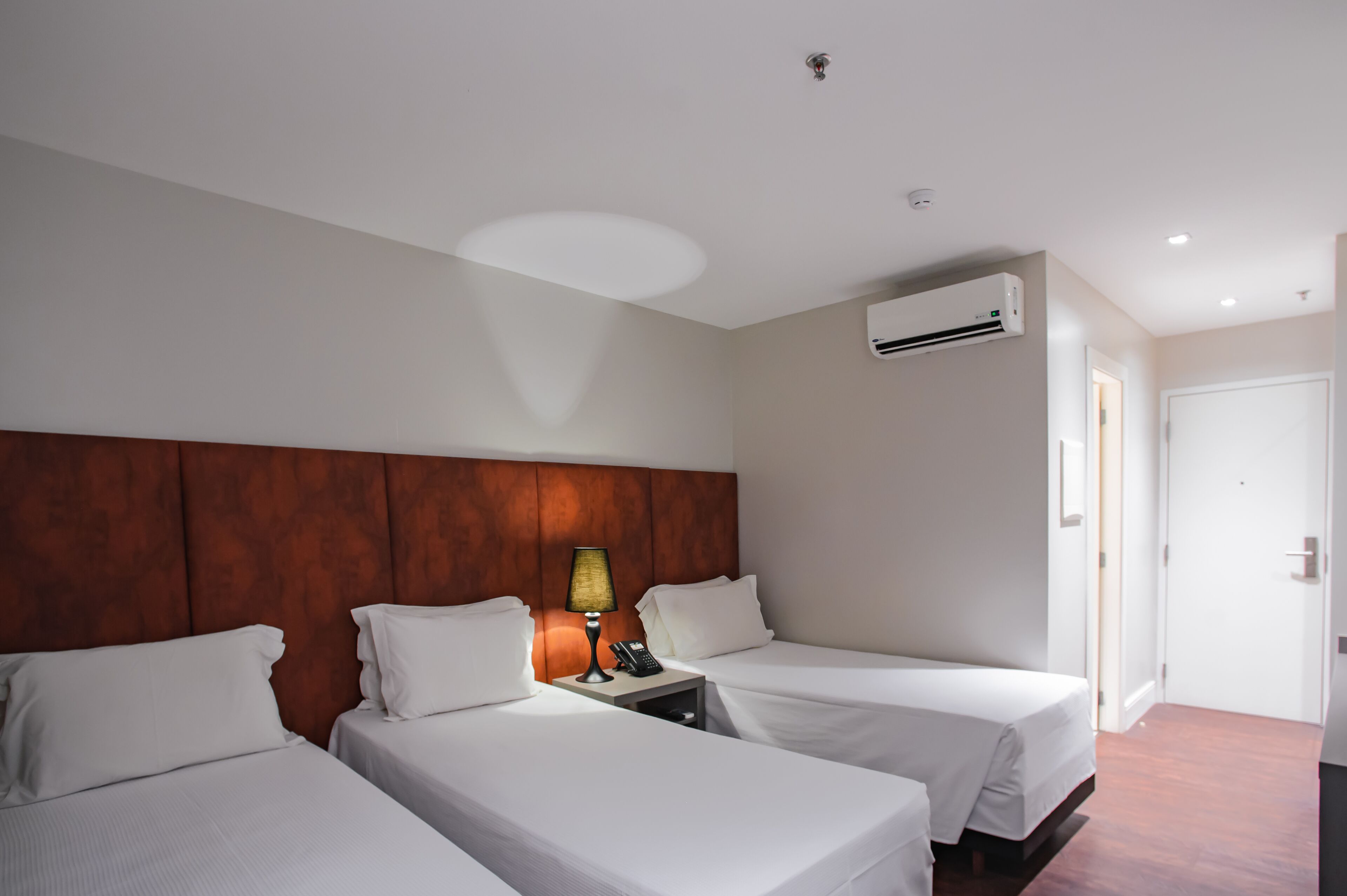 A hotel room with two single beds, a wooden headboard, a bedside table with a lamp, a phone, and an air conditioning unit.