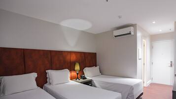 A hotel room with two single beds, a wooden headboard, a bedside table with a lamp, a phone, and an air conditioning unit.