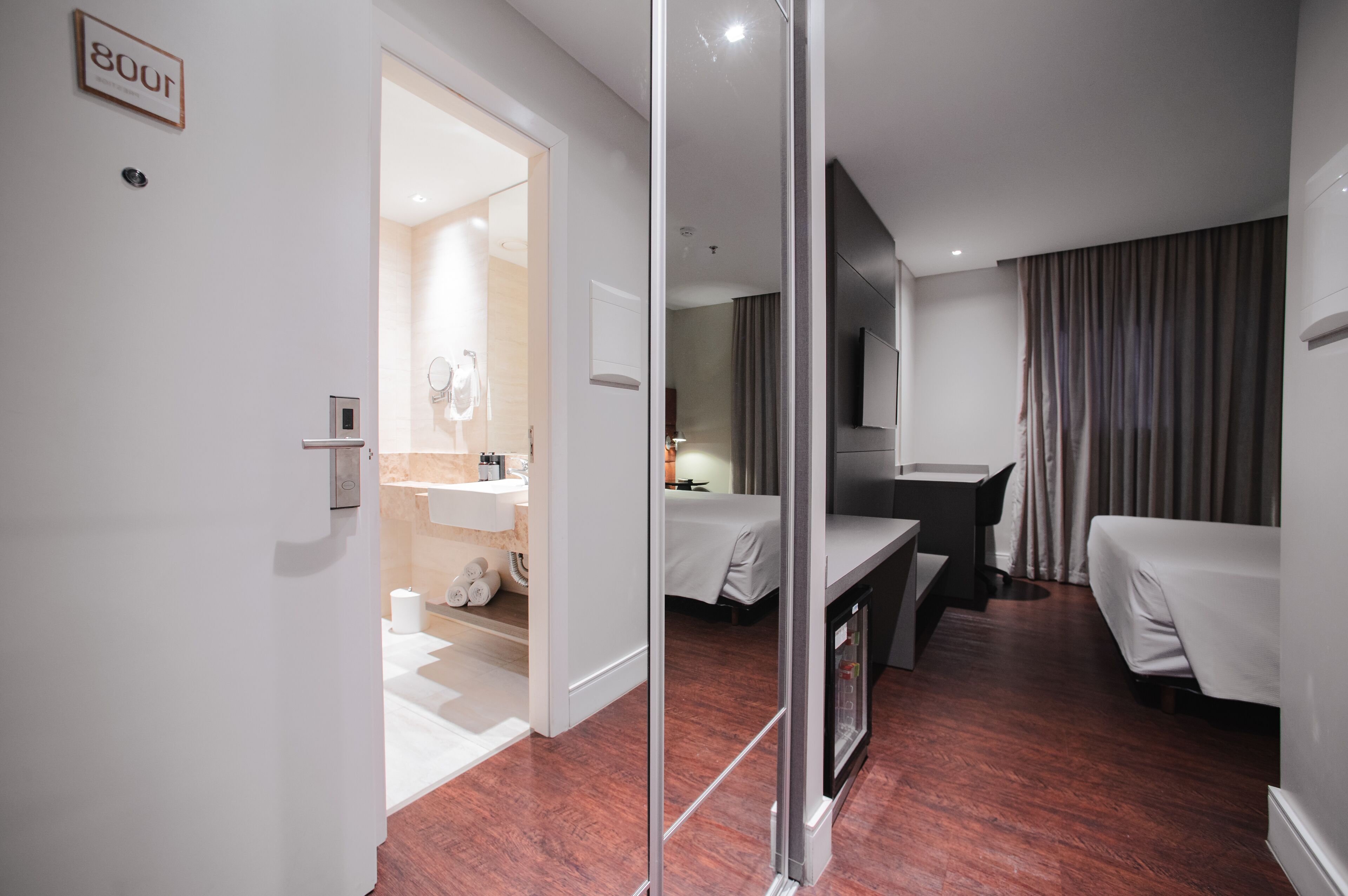 A modern hotel room with a bed, a desk, a mirror, and a bathroom visible through an open door.