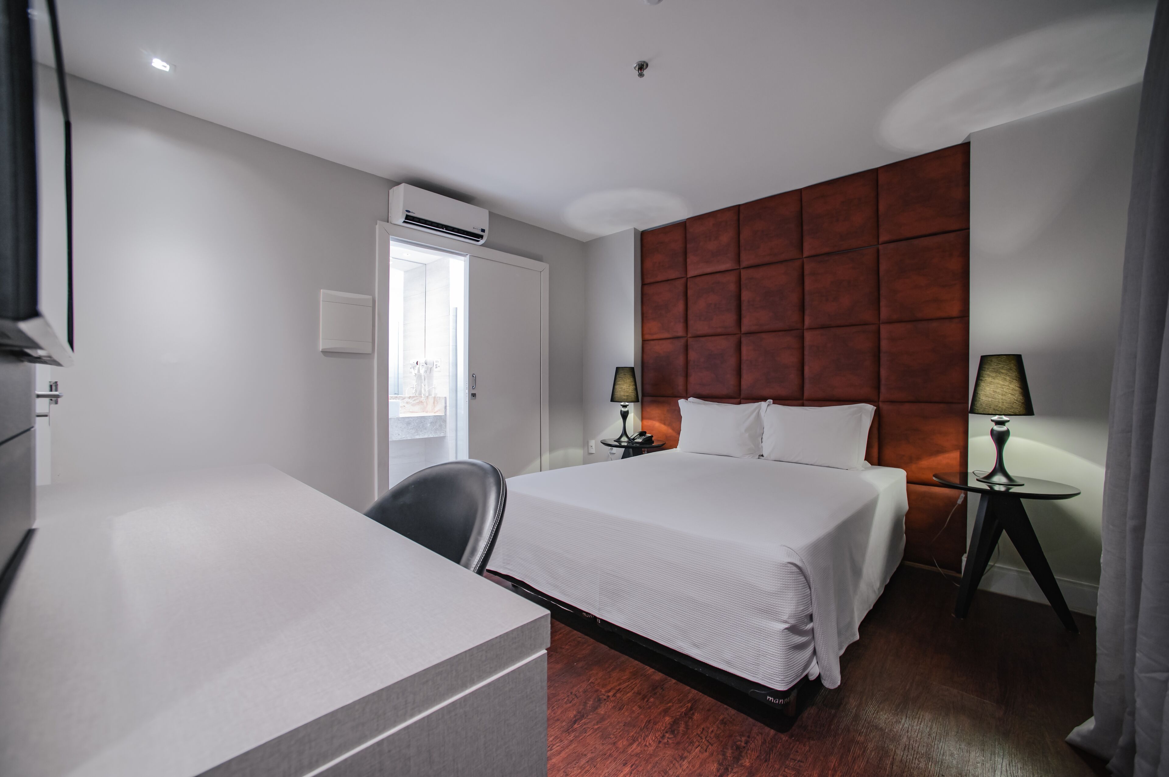 Habitación de hotel con cama, escritorio, silla, dos lámparas, aire acondicionado y ventana.