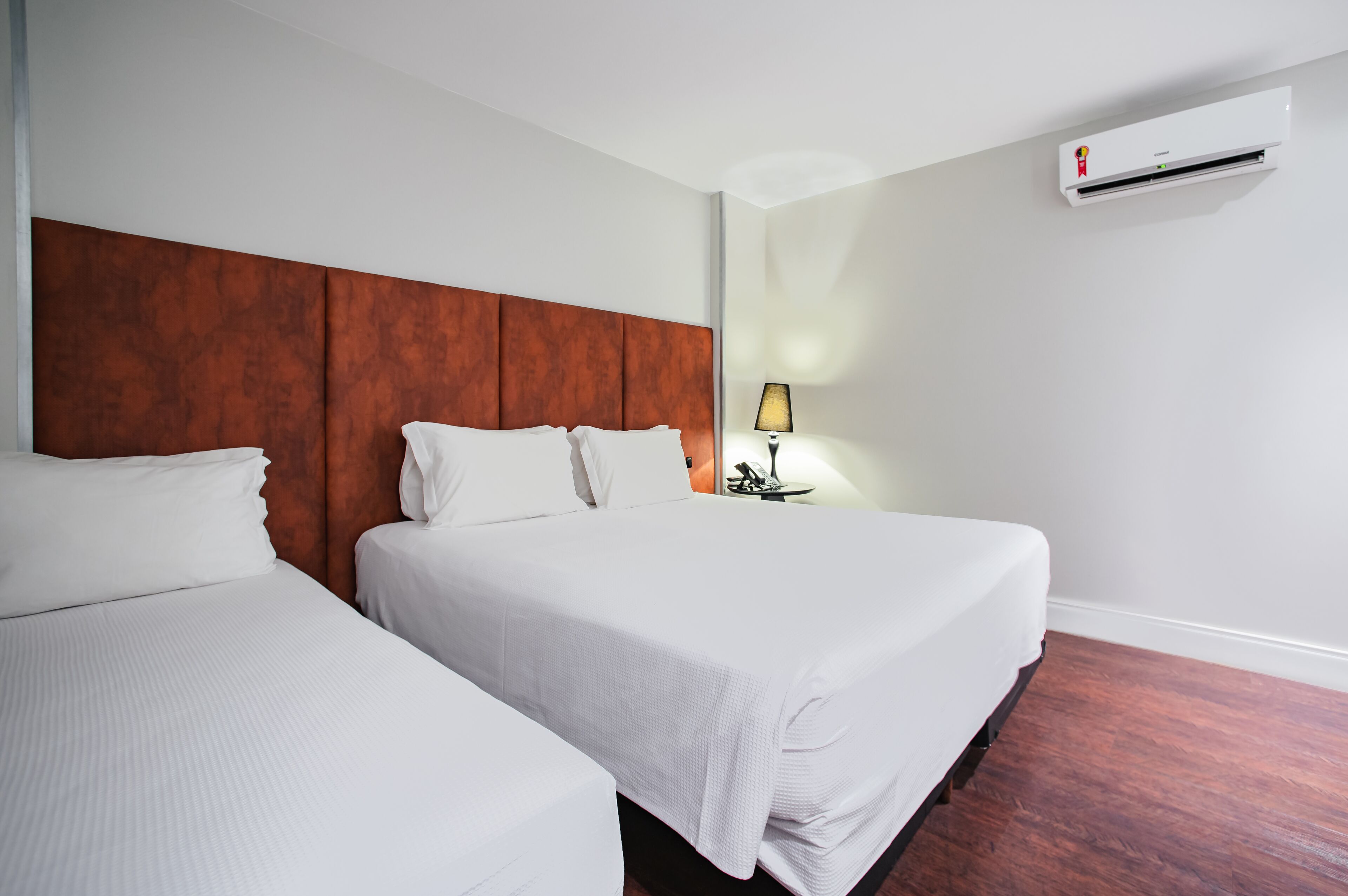Quarto de hotel com duas camas, cabeceira de madeira, mesa de cabeceira com abajur e ar condicionado.