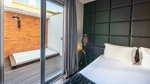 Ett modernt hotellrum med en stor säng, en mörk sänggavel och en glasdörr som leder ut till ett utomhusområde med ett trädäck och en mur av tegelsten.