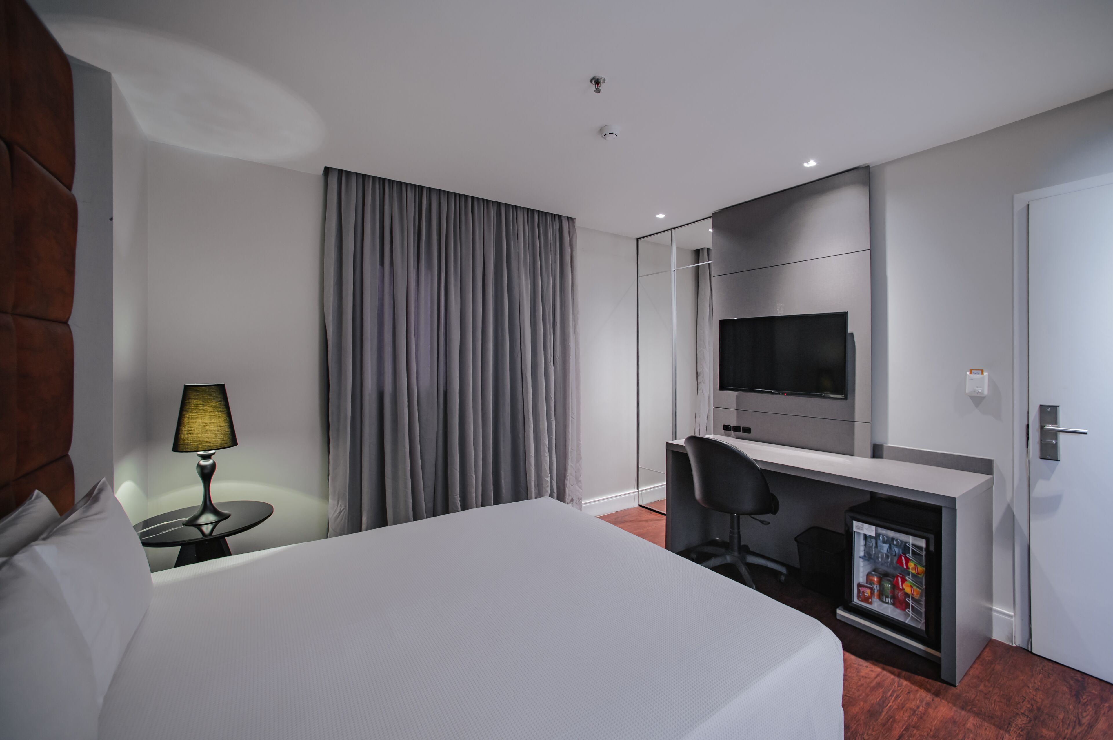 Habitación de hotel con cama, escritorio con silla, televisión, lámpara y minibar.