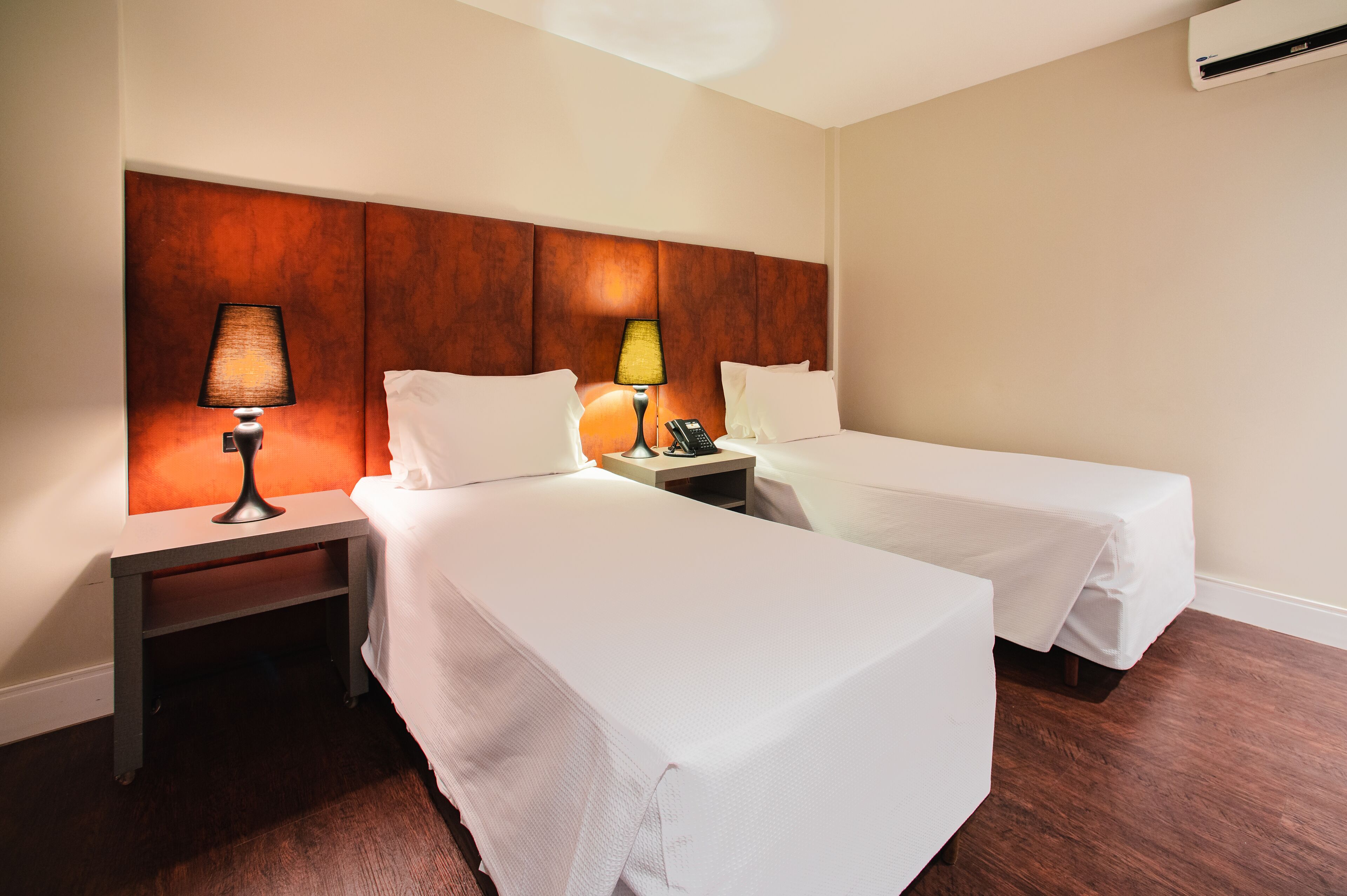 A hotel room with two single beds, a nightstand with a lamp, a phone, and an air conditioning unit on the wall.
