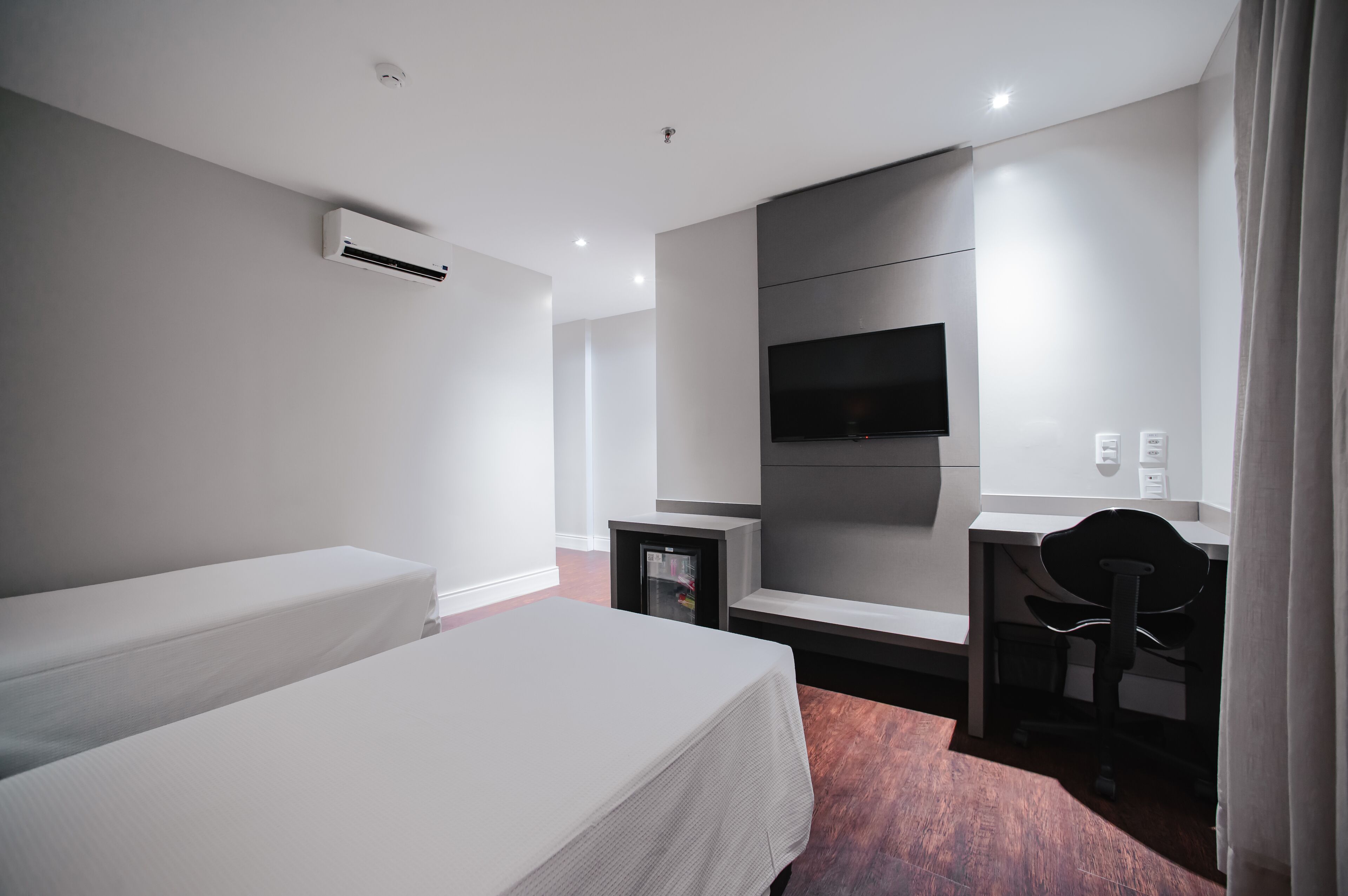 Quarto de hotel com uma cama, escrivaninha, cadeira, televisão e ar condicionado.