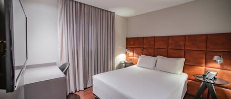Une chambre d’hôtel avec un grand lit, un bureau, une chaise, une table de chevet avec une lampe et un rideau.