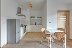 Una cocina compacta con refrigerador, estufa y gabinetes blancos. Hay sillas de mimbre y una mesa blanca.