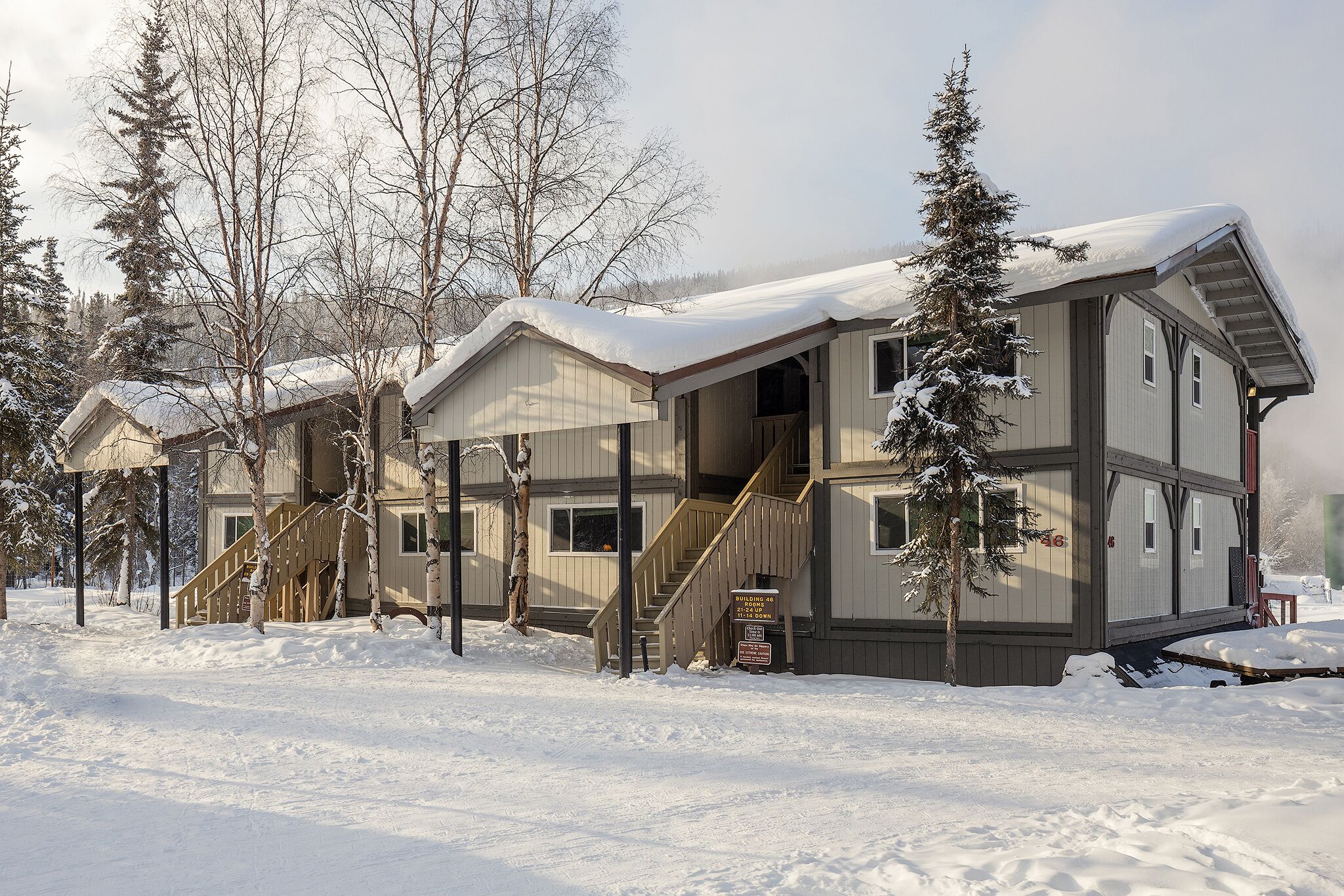 A multi-story lodge with a covered outdoor area, wooden stairs, and snow-covered surroundings.