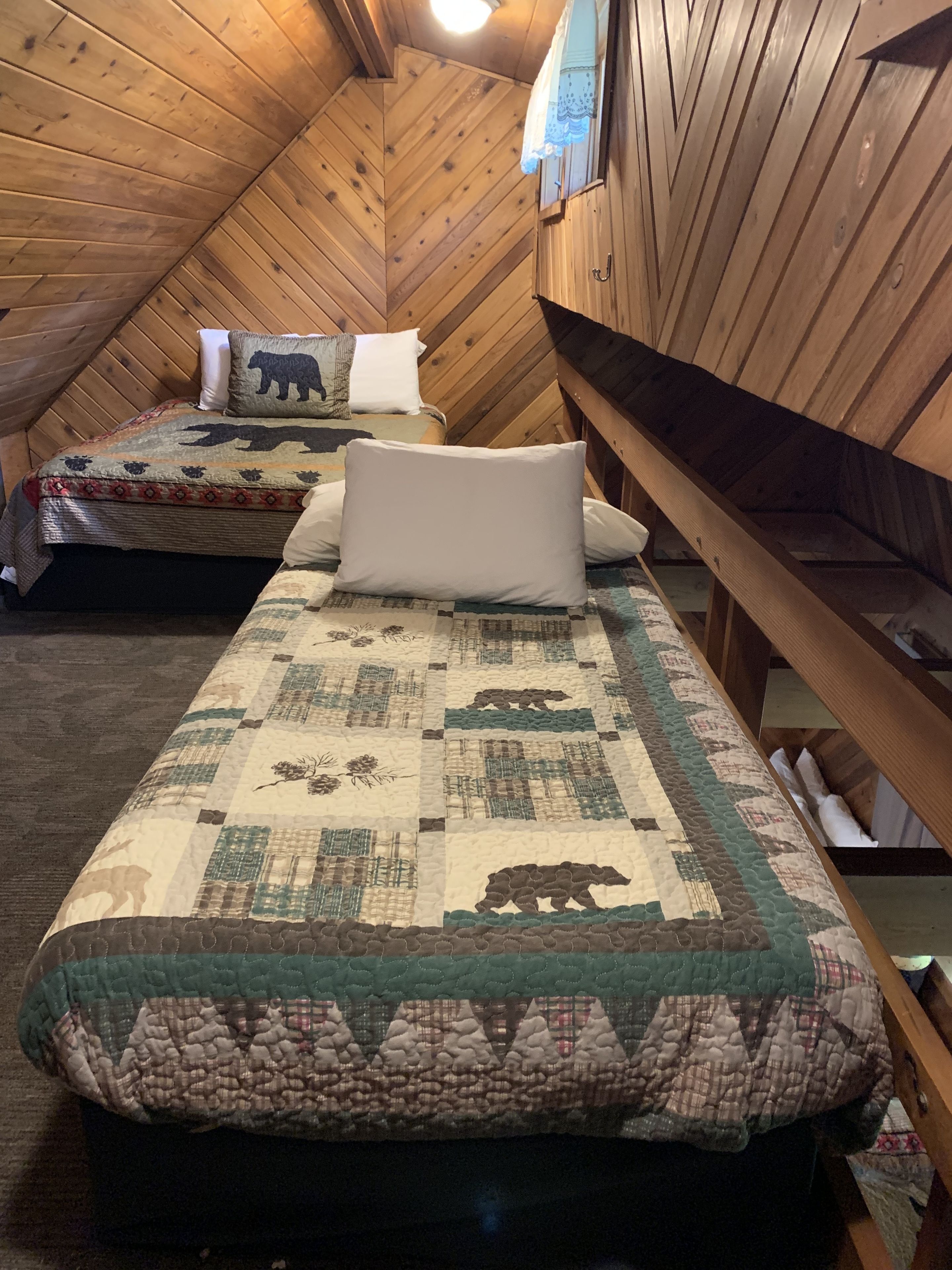 Ein gemütliches Schlafzimmer im Dachgeschoss mit Holzwänden und schrägen Decken. Es gibt zwei Betten mit gemusterten Bettdecken und Kissen, eines mit Bärenmotiv.