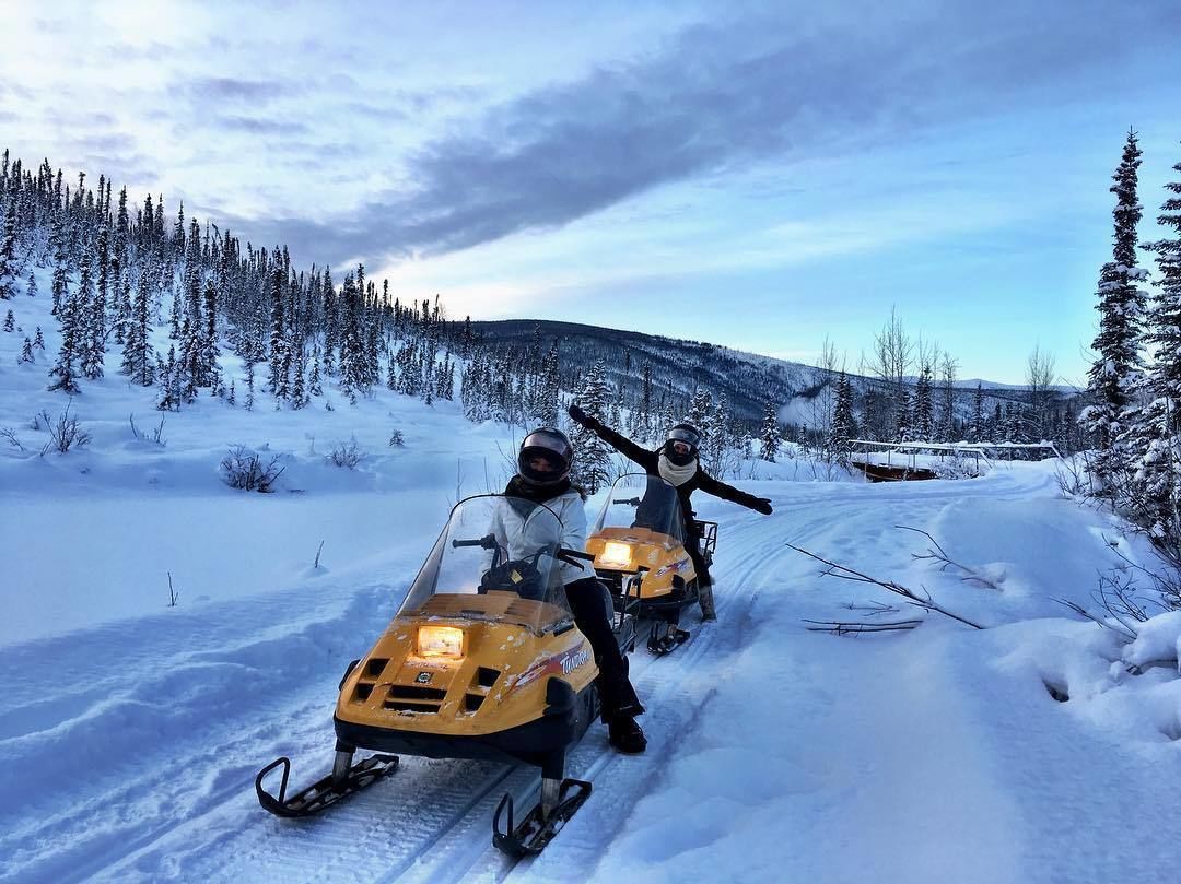 Two people on snowmobiles on a snowy road with trees and hills in the background.