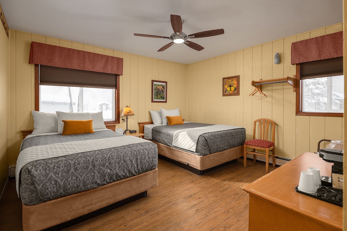 A hotel room with two beds, a ceiling fan, wooden flooring, a desk with a chair, and a window with a view.