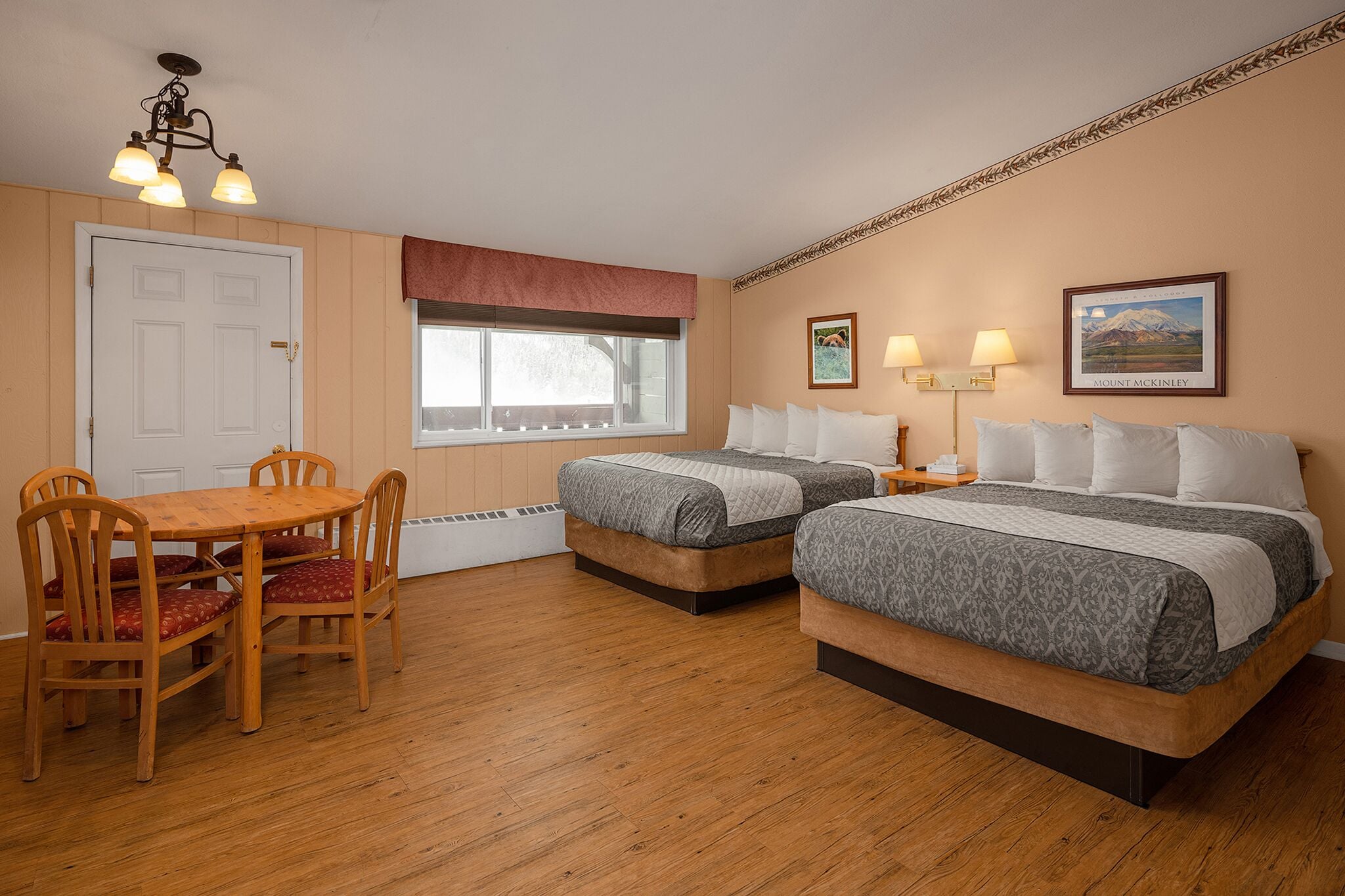 A room with two beds, a wooden dining table with chairs, a window with a view, and a ceiling light fixture.