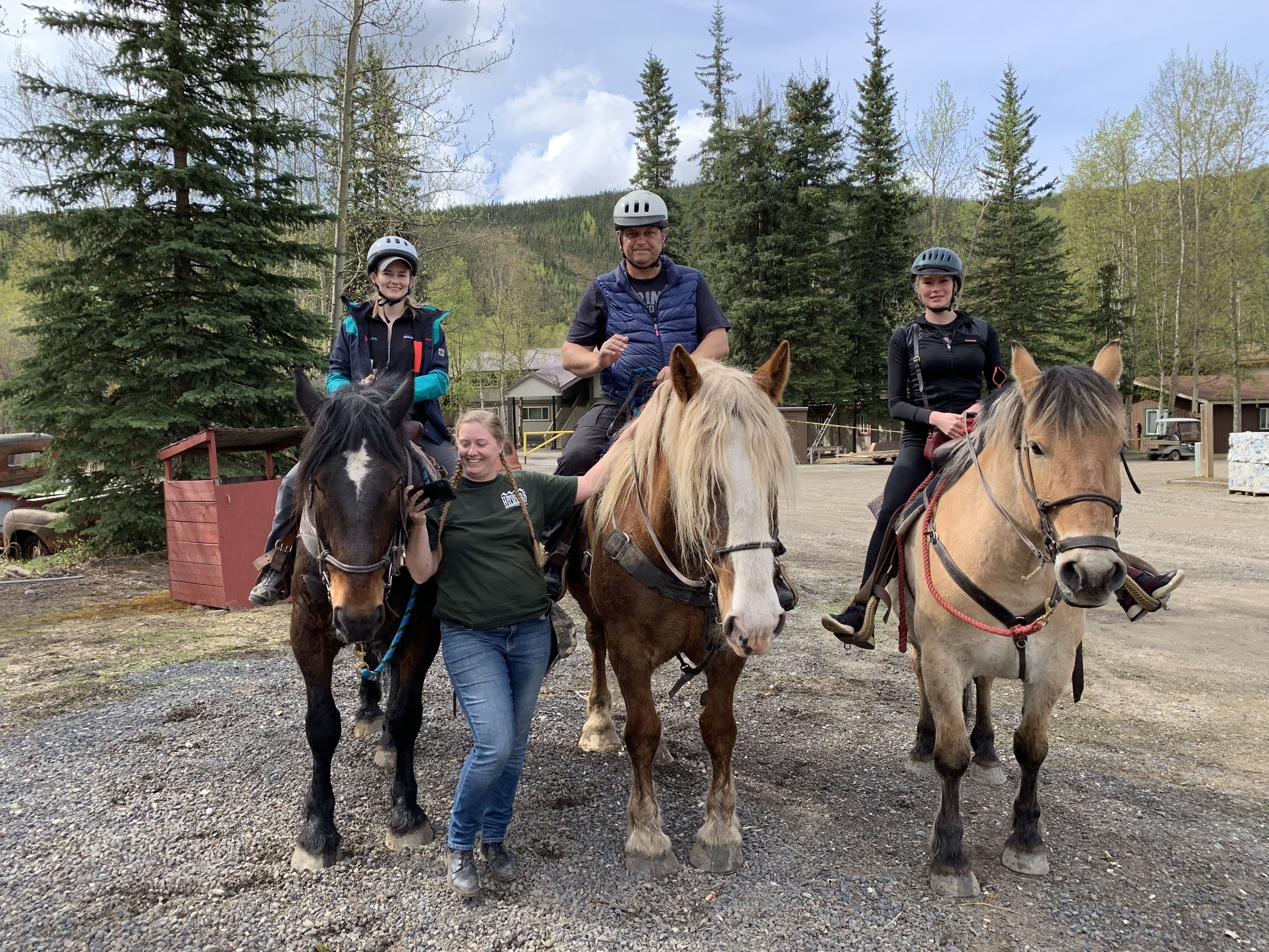 Three people on horseback in an outdoor setting with trees and a gravel path.