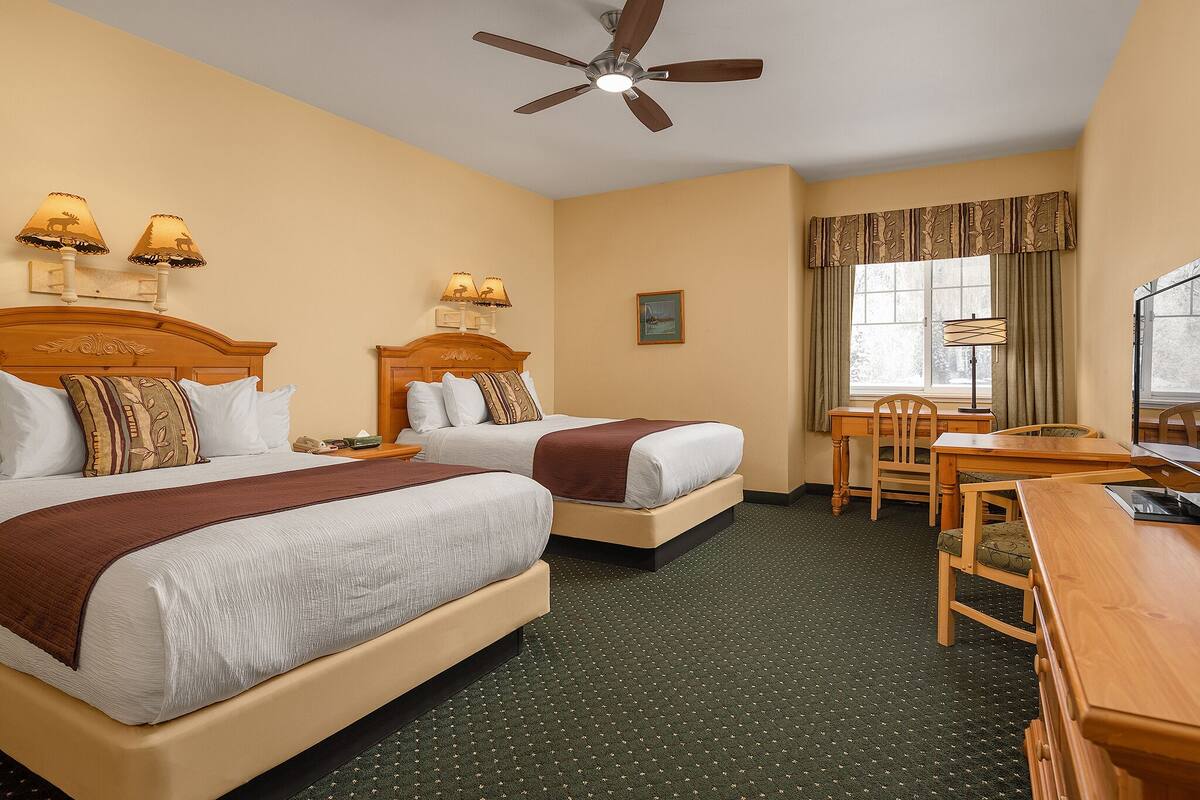 A hotel room with two beds, a desk, a chair, a ceiling fan, and a window with curtains.