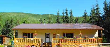 A two-story lodge with a wooden deck, a front porch with steps, and a mountain backdrop.