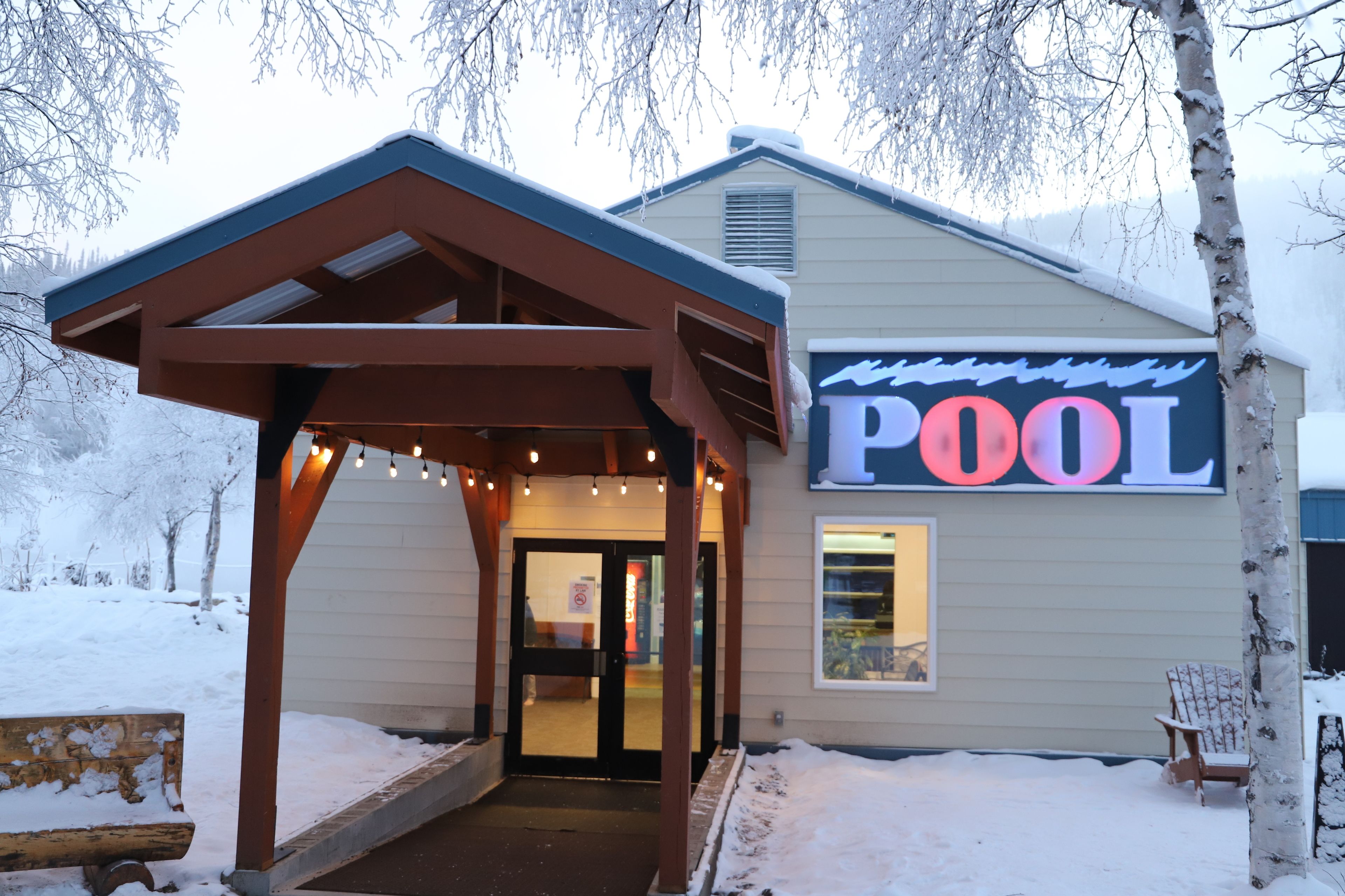A building with a 'POOL' sign, a covered entrance, and a bench.