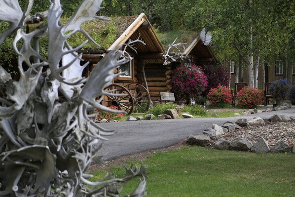A rustic log cabin with a wooden wheelbarrow in front, surrounded by greenery and flowering plants.
